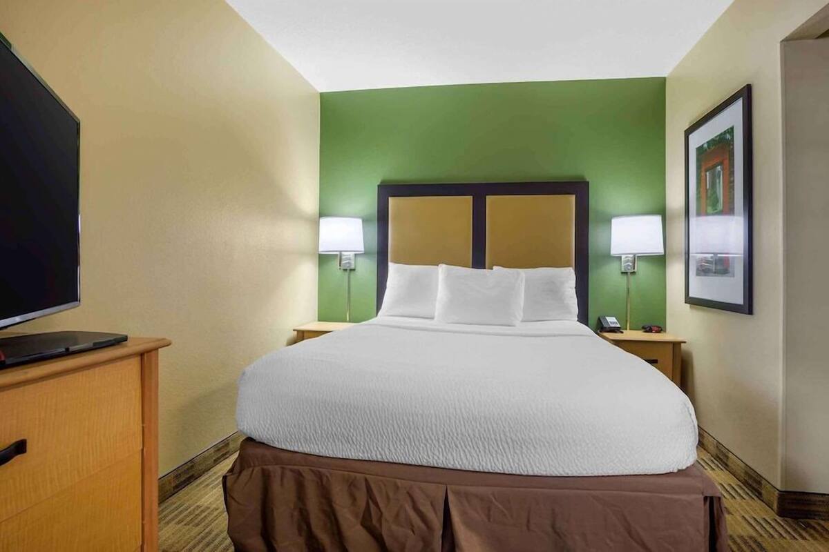 A hotel room with a large bed, two bedside lamps, a flat-screen TV, and a wooden nightstand.