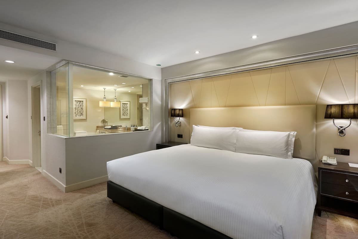 A modern hotel room with a large bed, a bedside table, a nightstand, a glass table, and a wall-mounted lamp.