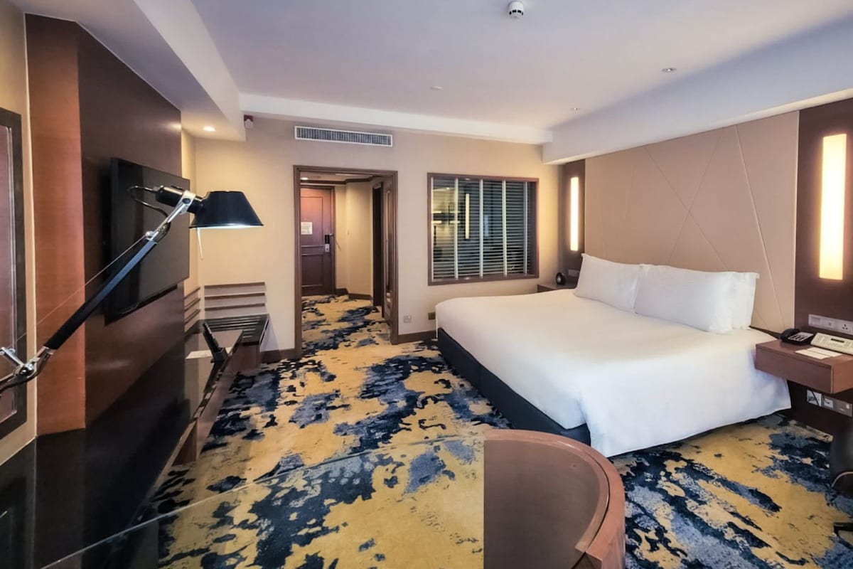 A hotel room with a large bed, a desk, a chair, a TV, and a window with blinds.