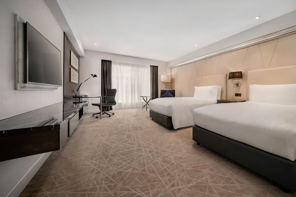 A hotel room with two beds, a desk with a computer, a TV, and a large window with curtains.