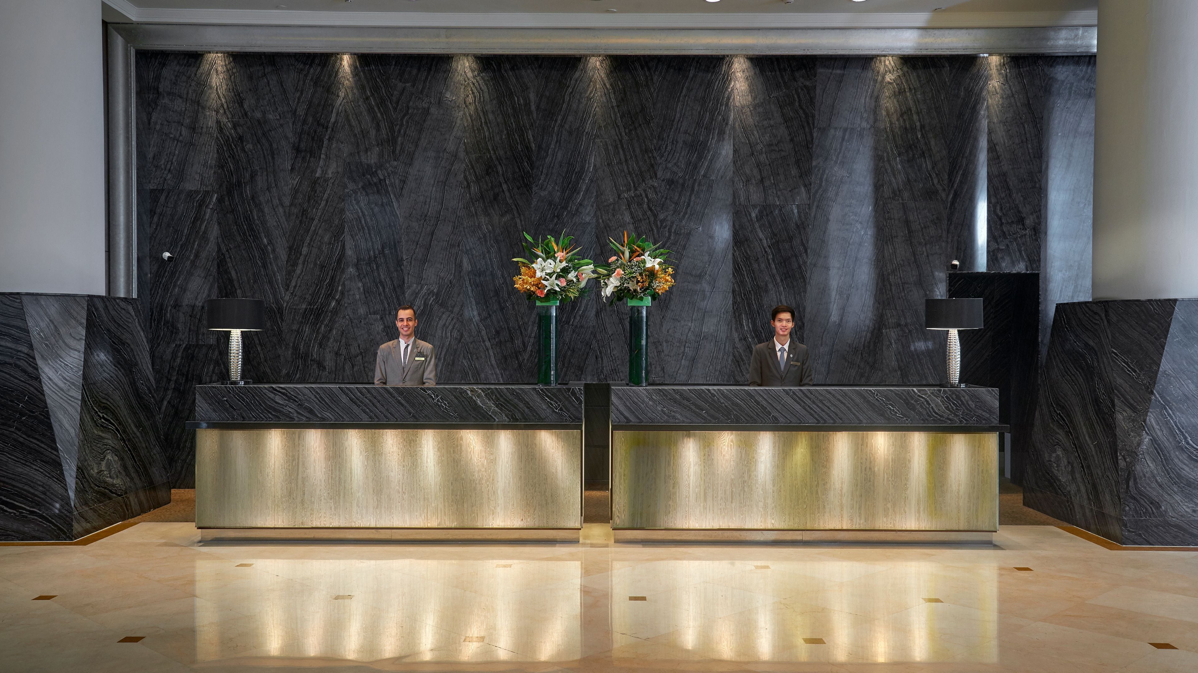 Un mostrador de recepción con dos recepcionistas, una pared de mármol y dos arreglos florales.