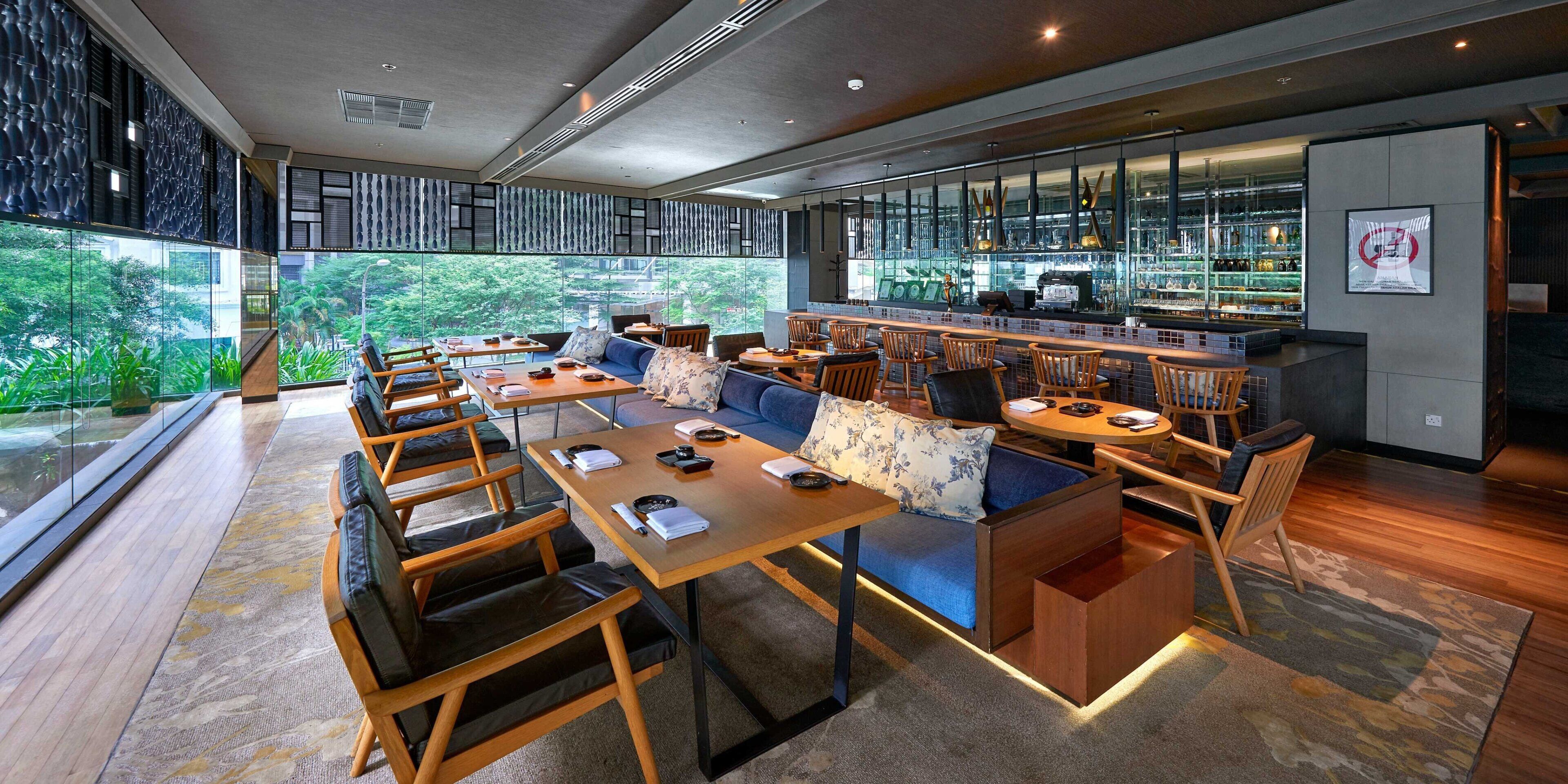 A modern restaurant with a bar area, seating arrangements, and large windows offering a view of greenery.