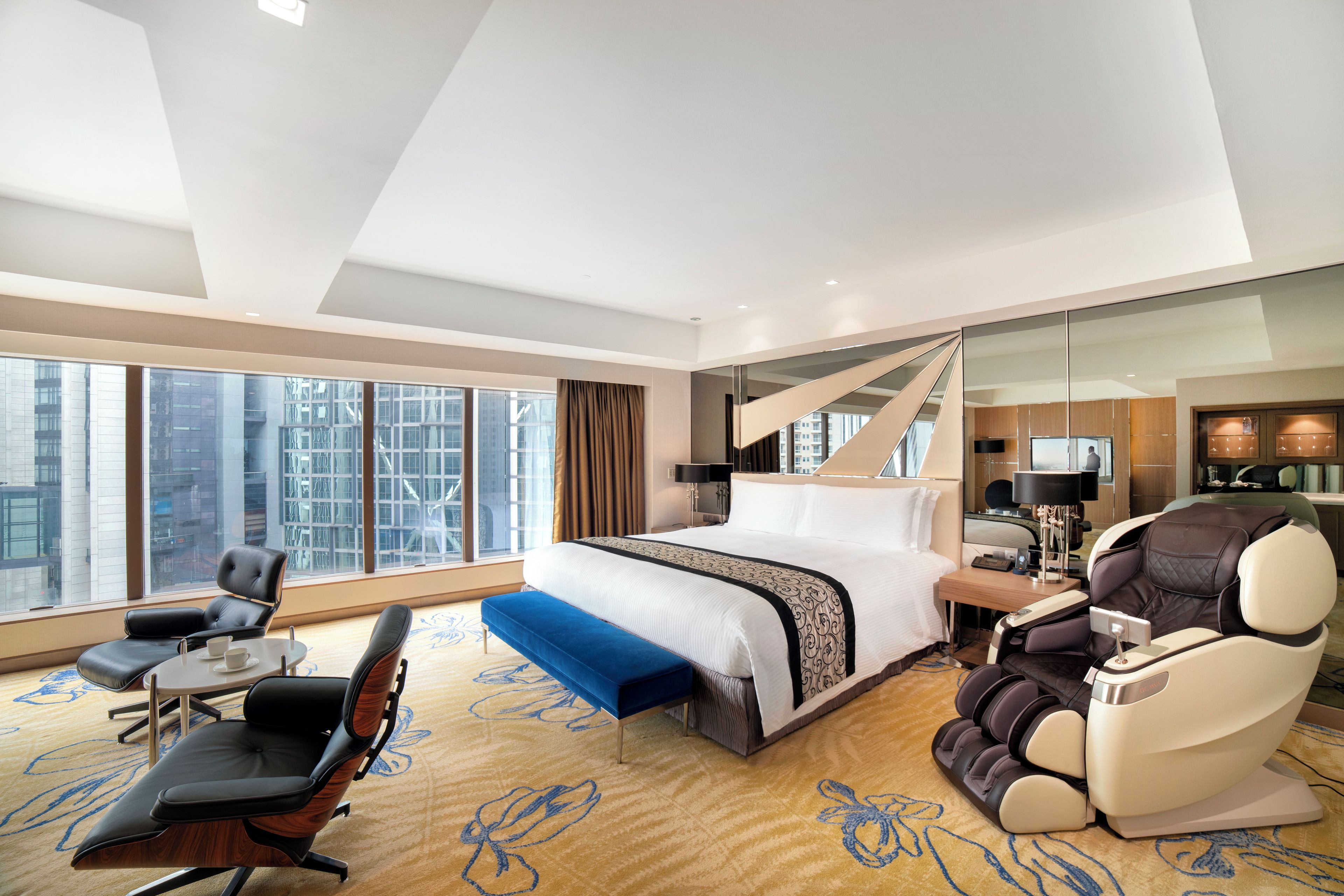A modern hotel room with a large bed, a massage chair, a desk with a lamp, and a view of the city through large windows.
