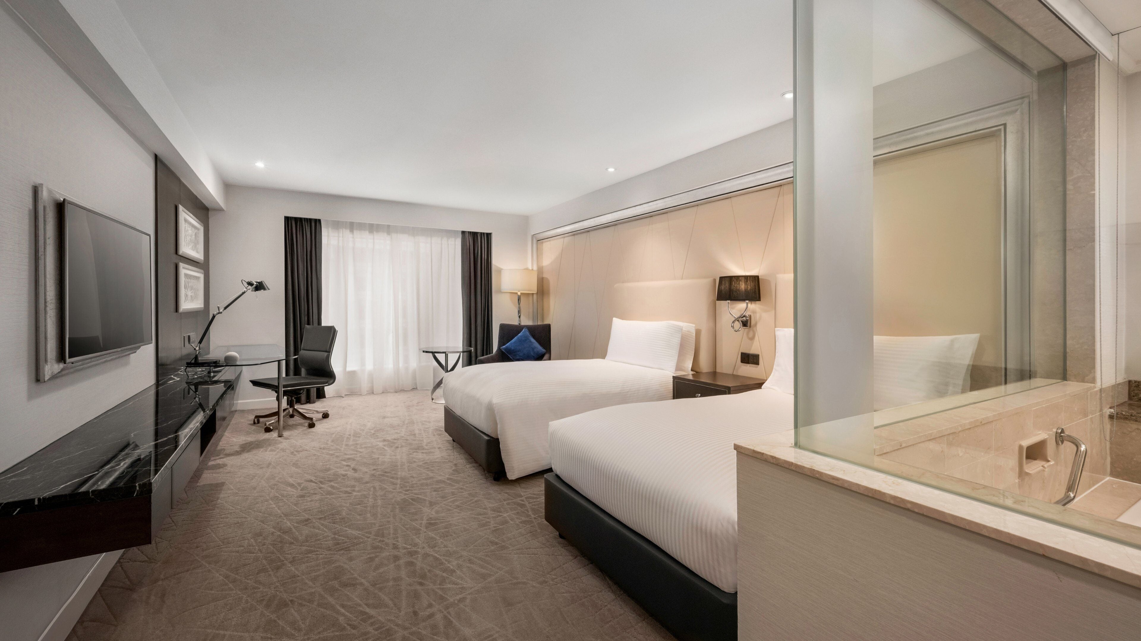 Habitación de hotel con dos camas, un escritorio con silla, televisión y baño con cabina de ducha de vidrio.