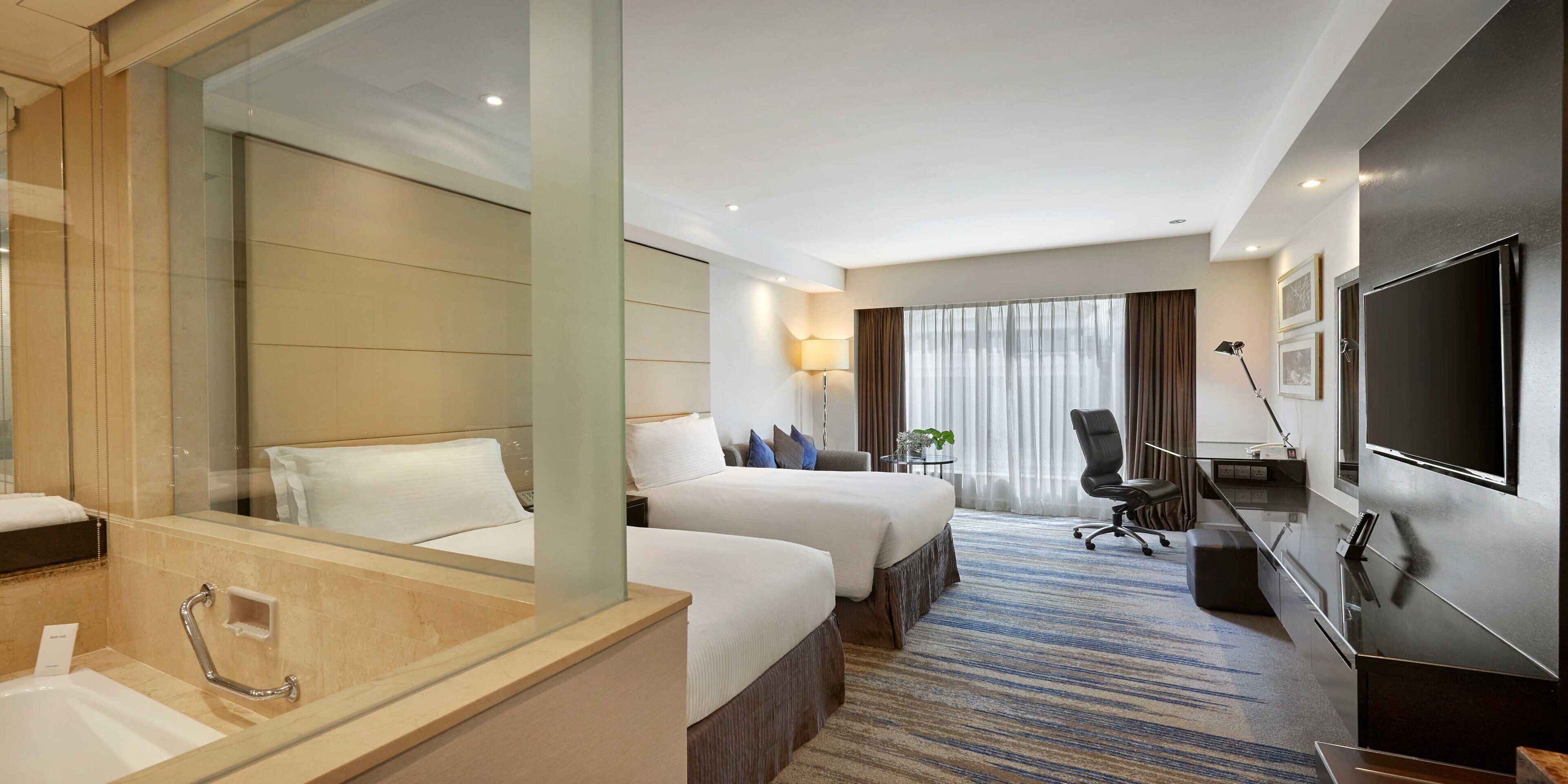 A hotel room with a large mirror, two beds, a desk with a chair, a TV, and a bathroom with a sink and mirror.