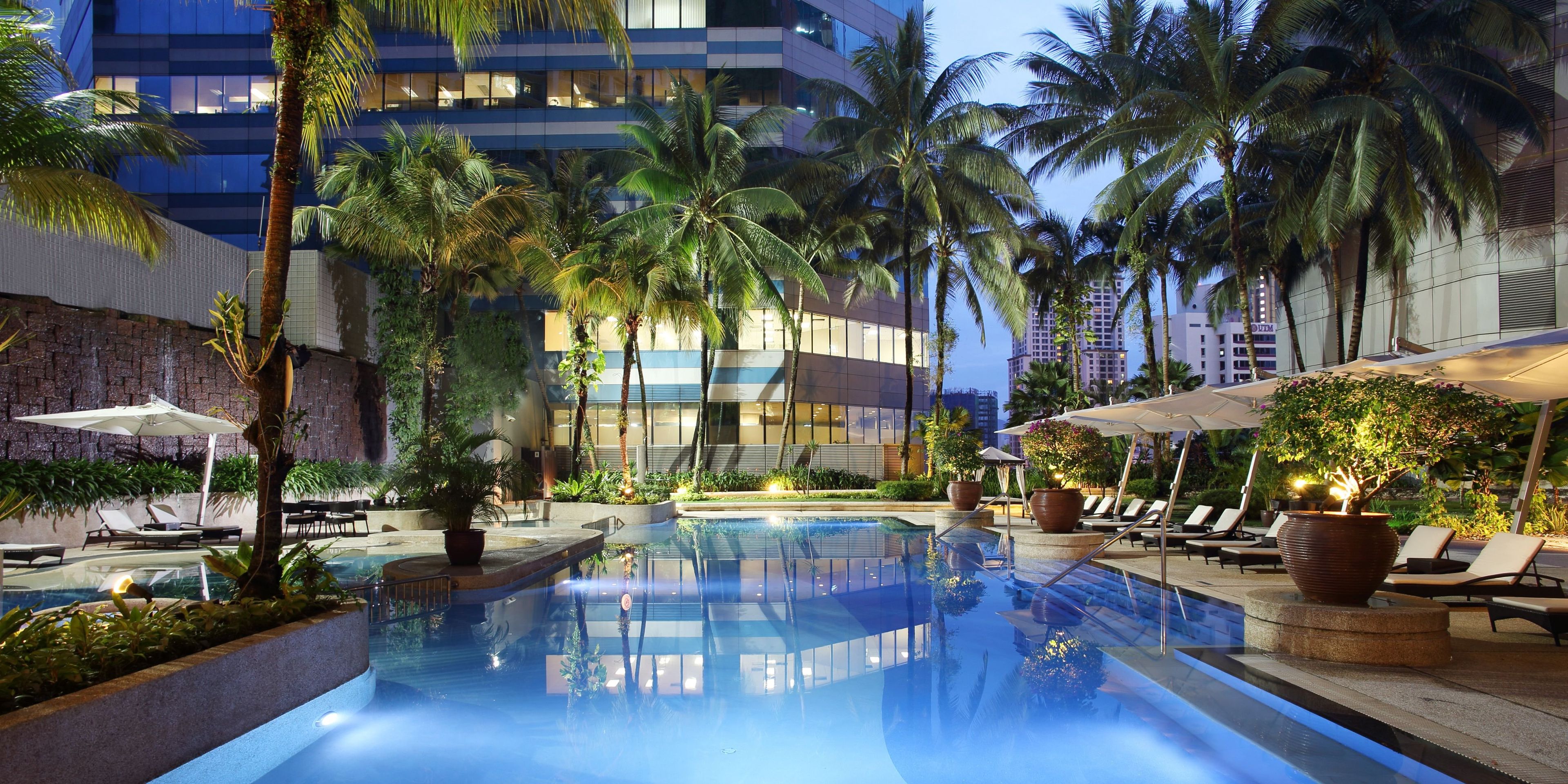 A pool area with lounge chairs and umbrellas, surrounded by palm trees and modern buildings.