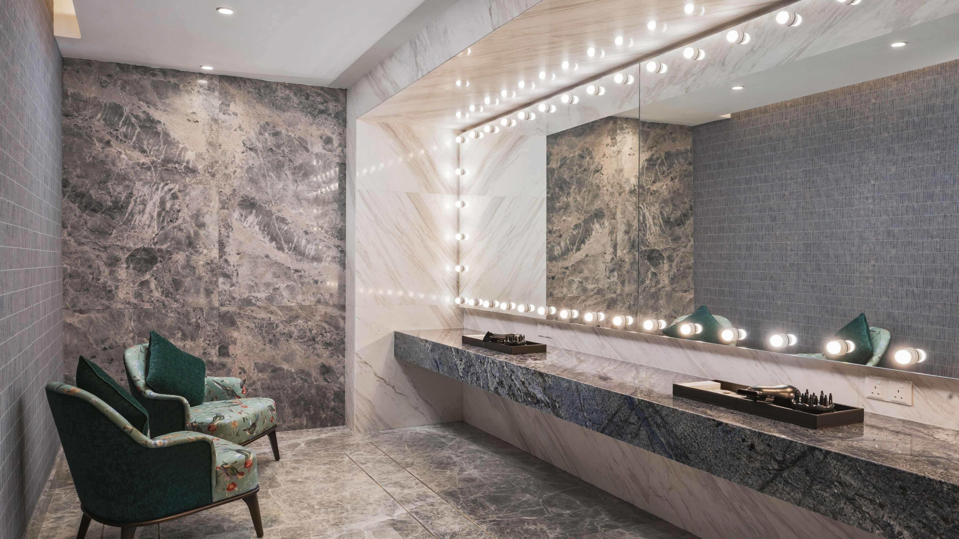Un baño con tocador de mármol, un espejo grande con iluminación y dos sillones tapizados en verde.