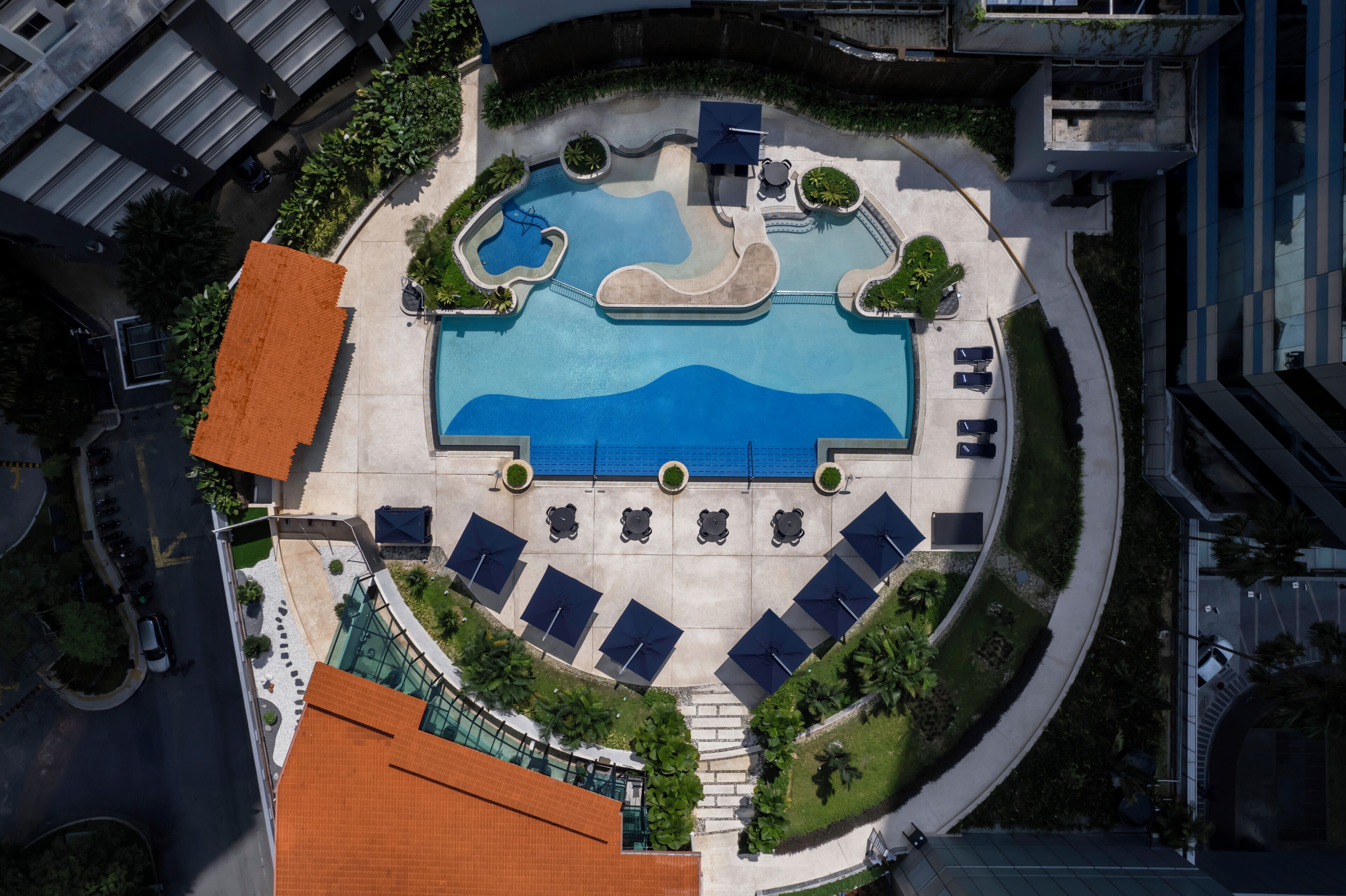 An aerial view of a hotel with a central swimming pool, sun loungers, and a landscaped area with trees and shrubs.
