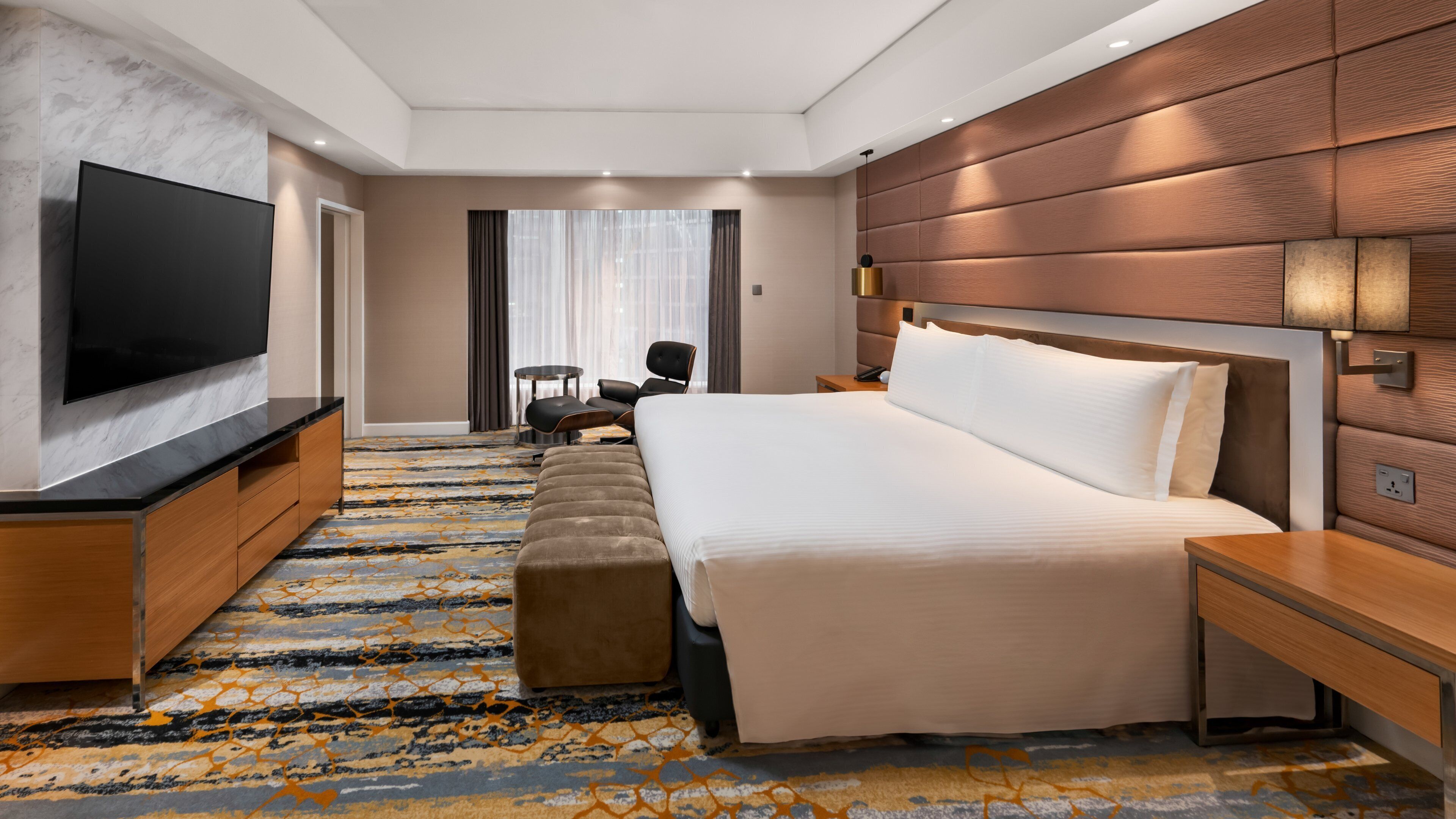 A modern hotel room with a large bed, a flat-screen TV, a wooden headboard, and a patterned carpet.