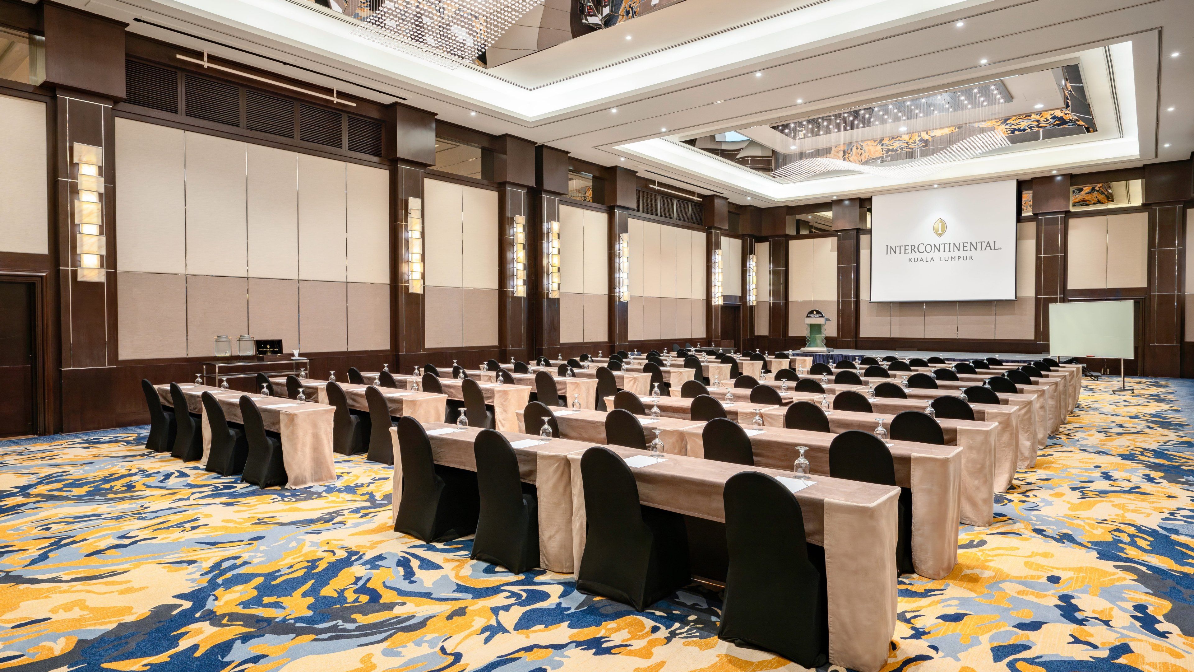A conference room with rows of tables and chairs, a large screen, and a patterned carpet.