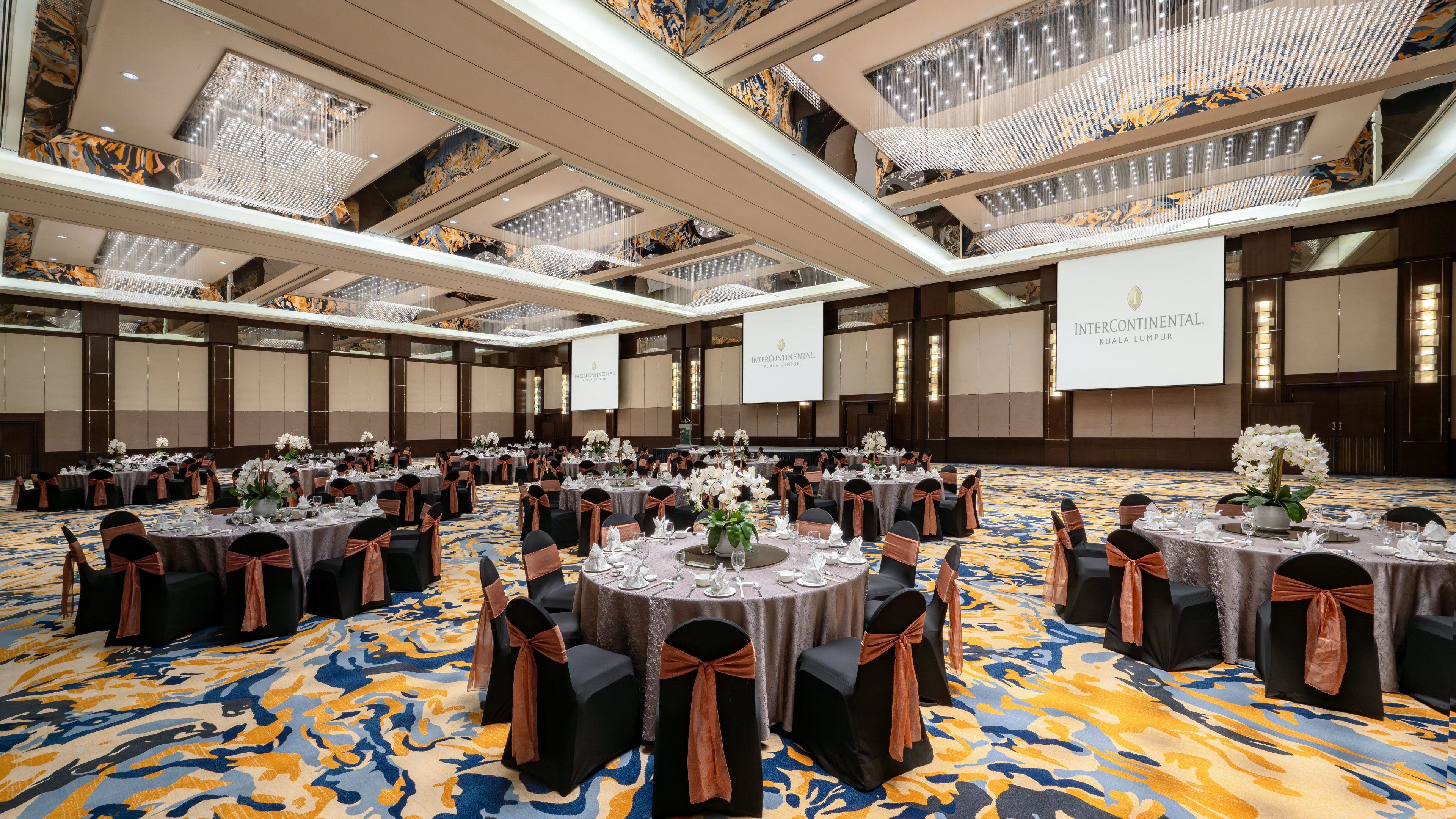 A spacious banquet hall with round tables, black chairs with brown sashes, and a patterned carpet.