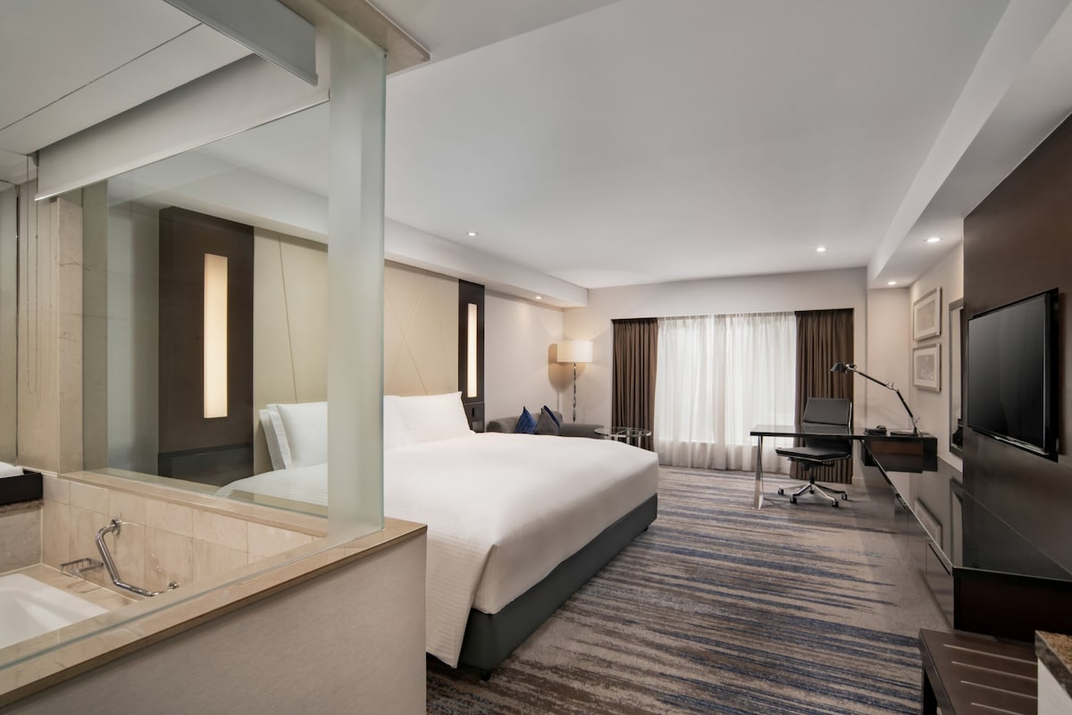 A hotel room with a large bed, a desk, a TV, and a bathroom with a glass-enclosed shower.