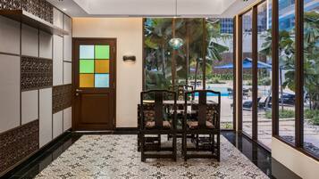 Un hall de hotel con piso de baldosas, una mesa de comedor de madera con sillas, una puerta decorativa y ventanas grandes con vistas a una zona de piscina.
