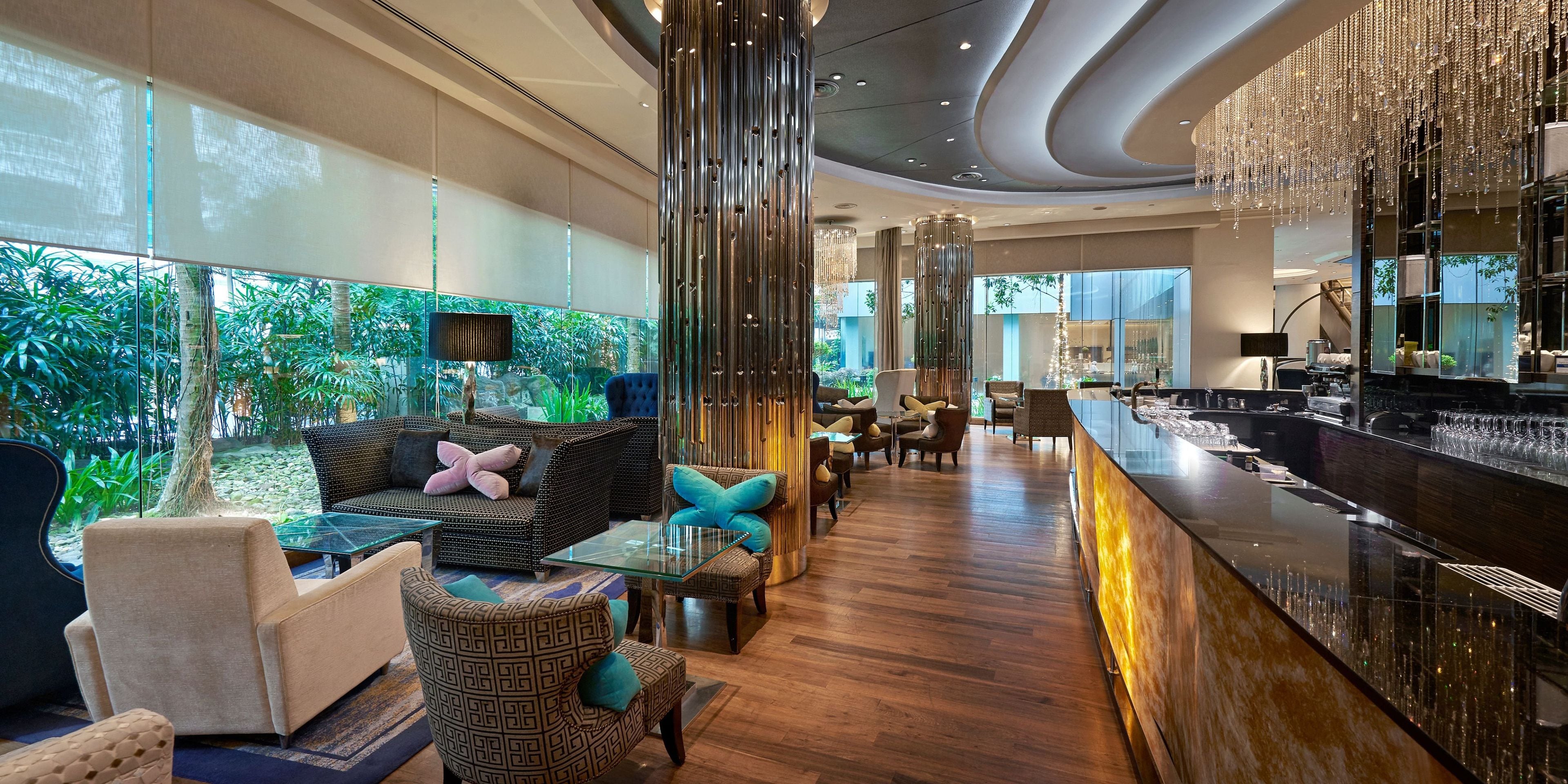 En moderne hotellobby med glasvæg, siddeområde, bar og udsigt til grønne områder.