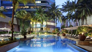 Ein Hotelpoolbereich mit Liegestühlen, Palmen und einem modernen Gebäude im Hintergrund.