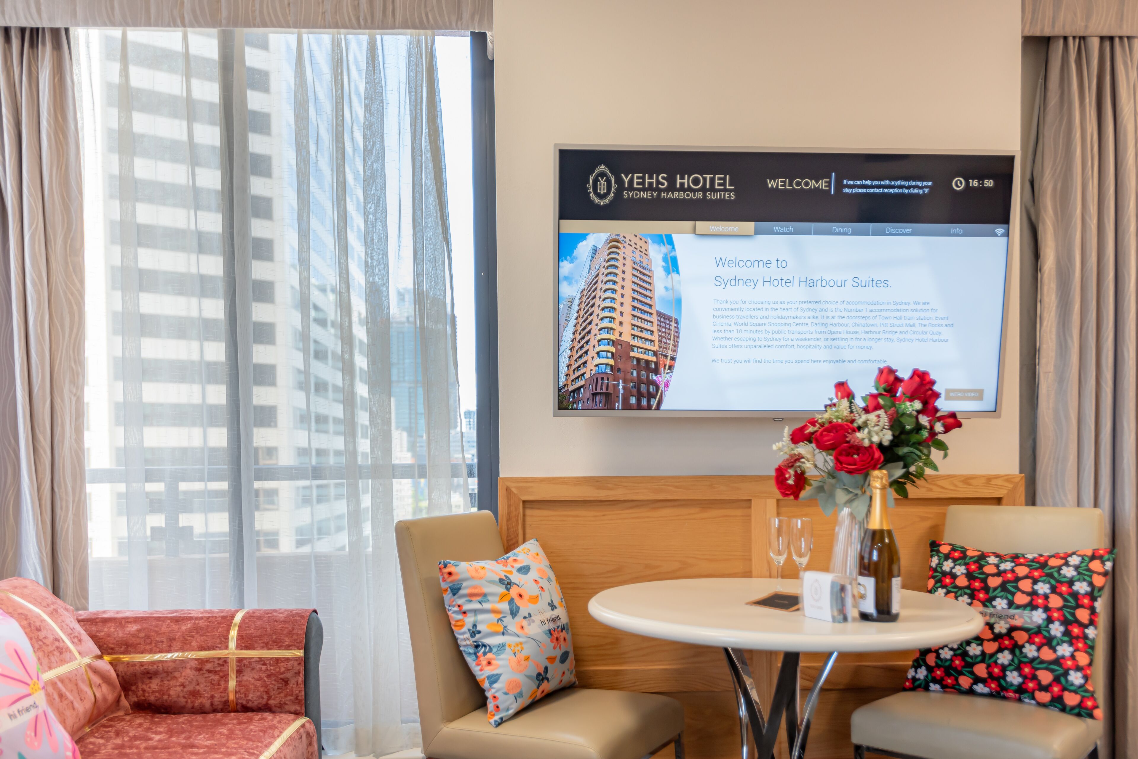 A hotel room with a large screen displaying the hotel's welcome page, a round table with a vase of flowers, and two armchairs with patterned cushions.