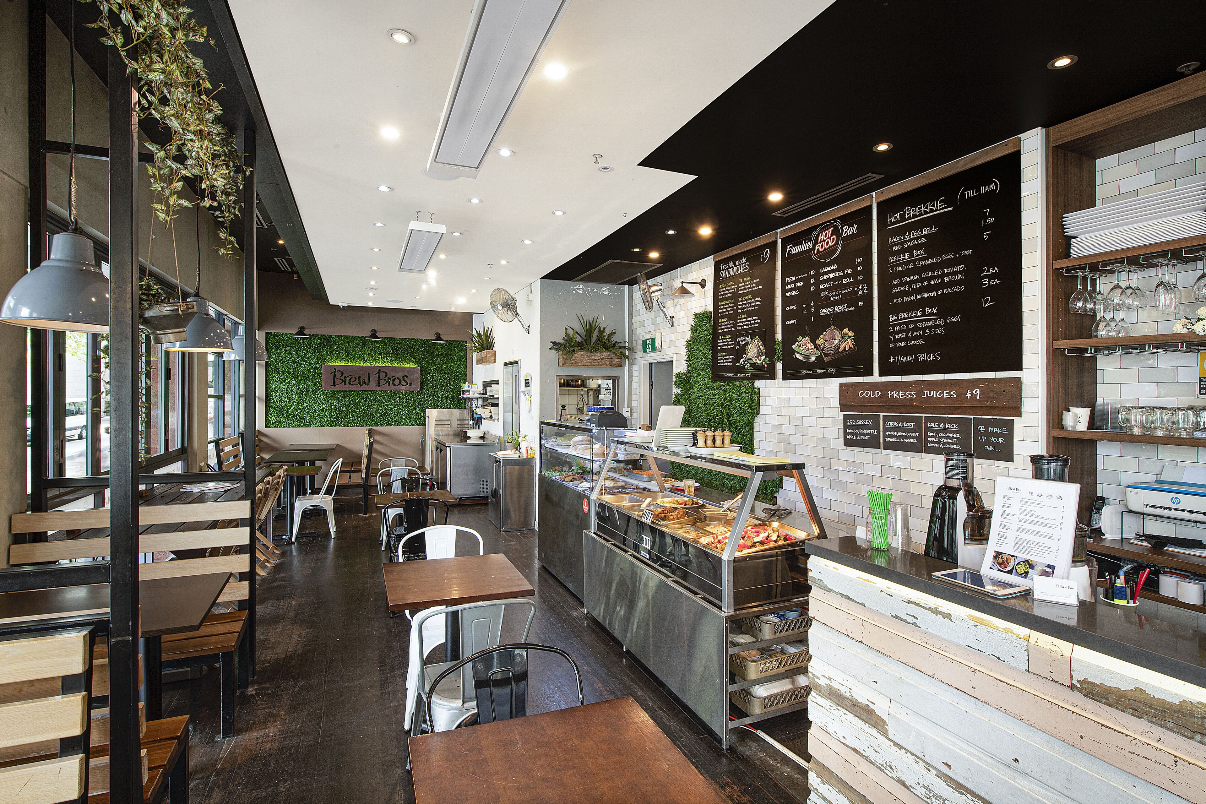 L’intérieur d’un café, avec des tables et des chaises en bois, un comptoir où est affiché le menu, et un mur végétal orné de plantes.
