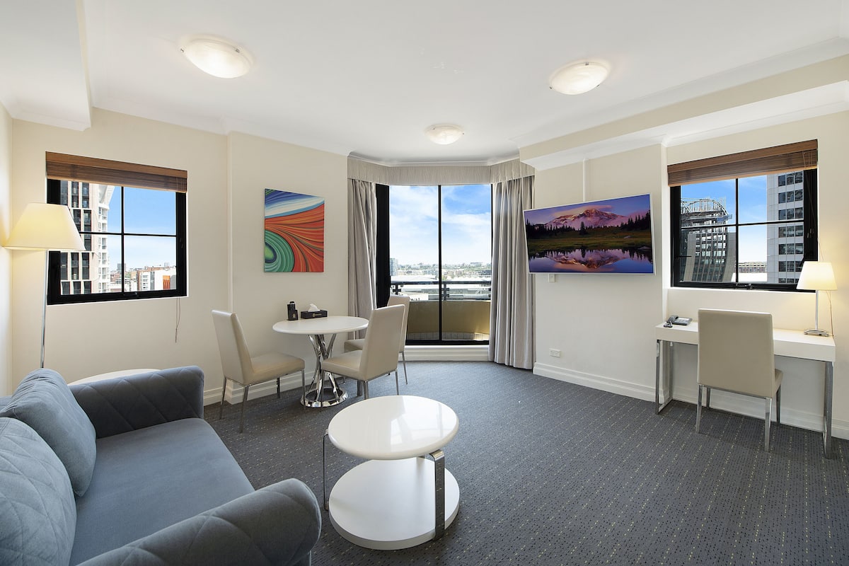 A hotel room with a gray sofa, a small round table, a desk with a chair, and large windows offering city views.