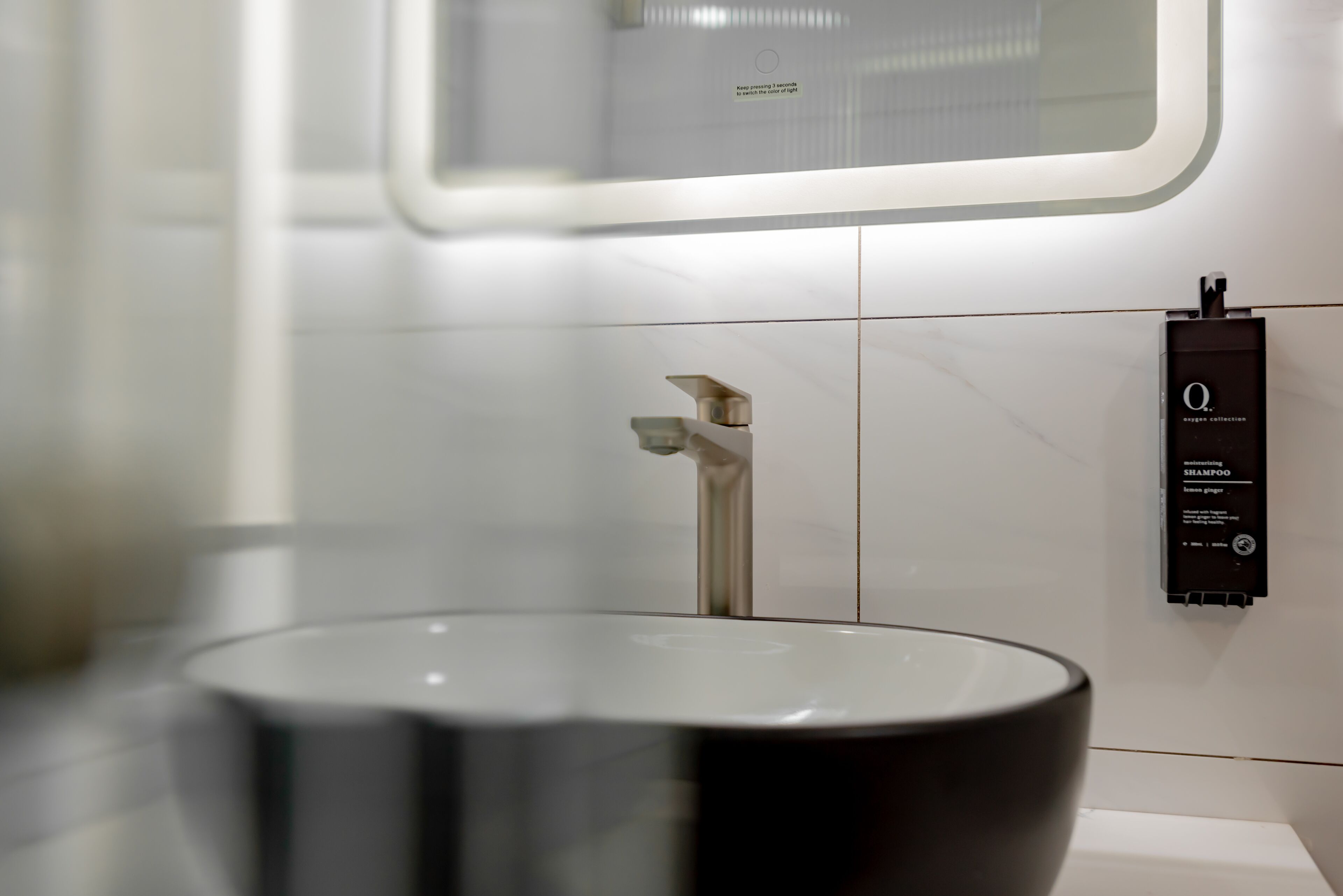A modern bathroom with a large, round, black sink and a sleek, metallic faucet. A black dispenser is mounted on the wall to the right of the sink.
