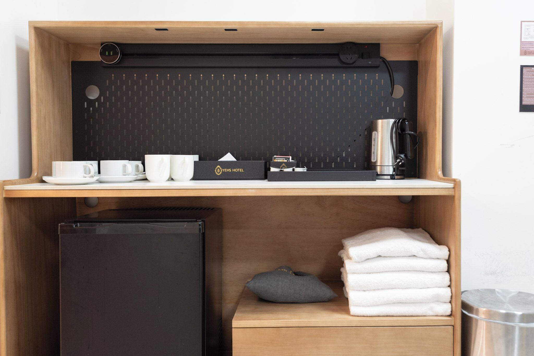 A compact hotel room with a wooden desk, a black refrigerator, a stack of white towels, and a small gray pillow.