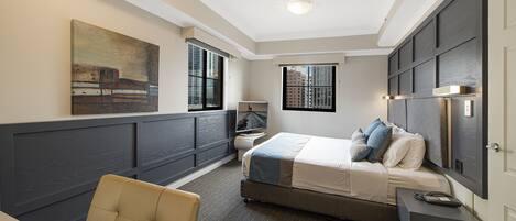 A hotel room with a bed, bedside table, chair, artwork on the wall, windows, and a view of buildings outside.