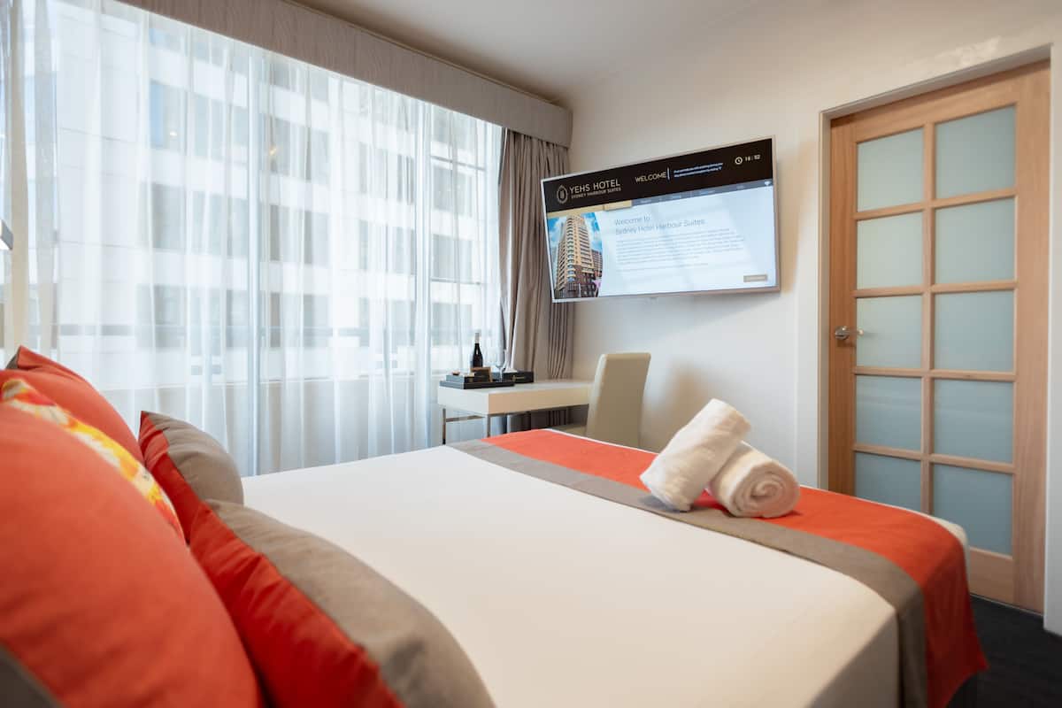 Ein Hotelzimmer mit Bett, Schreibtisch, Stuhl, einem Fernseher, der Hotelinformationen zeigt, und einem Fenster mit Vorhängen.