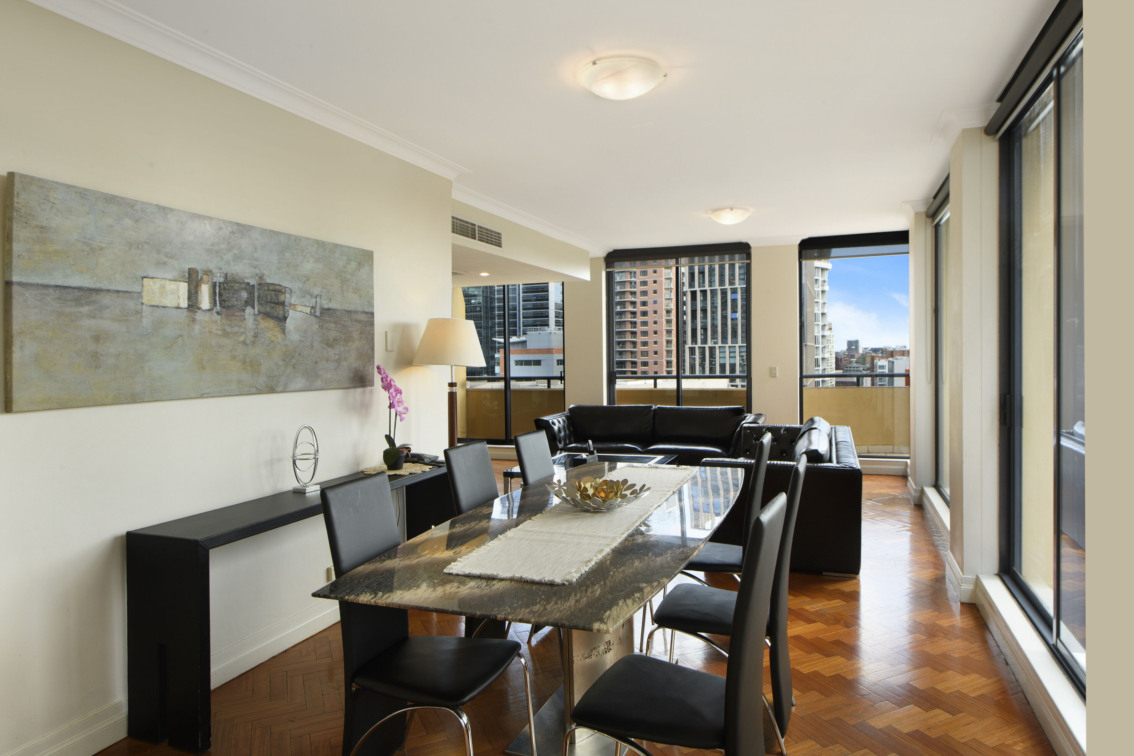 A modern dining area with a glass table, black chairs, and a large abstract painting on the wall.