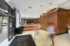 Un área de recepción de hotel con un diseño moderno, que incluye un mostrador de recepción, un felpudo negro con el nombre del hotel y un amplio ventanal con vistas a la calle.