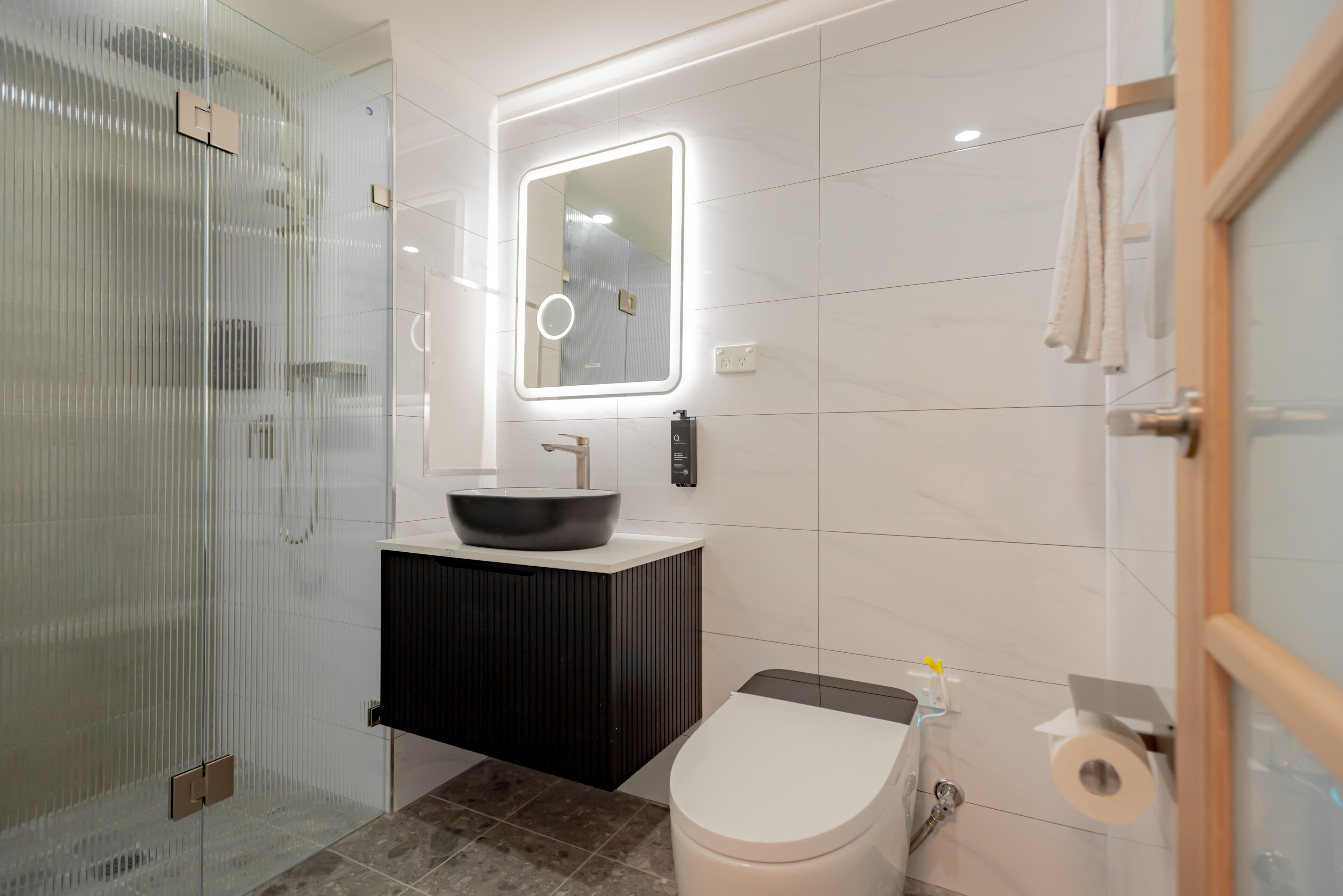 A modern bathroom with a glass shower enclosure, a black vanity with a vessel sink, and a toilet with a paper towel dispenser.