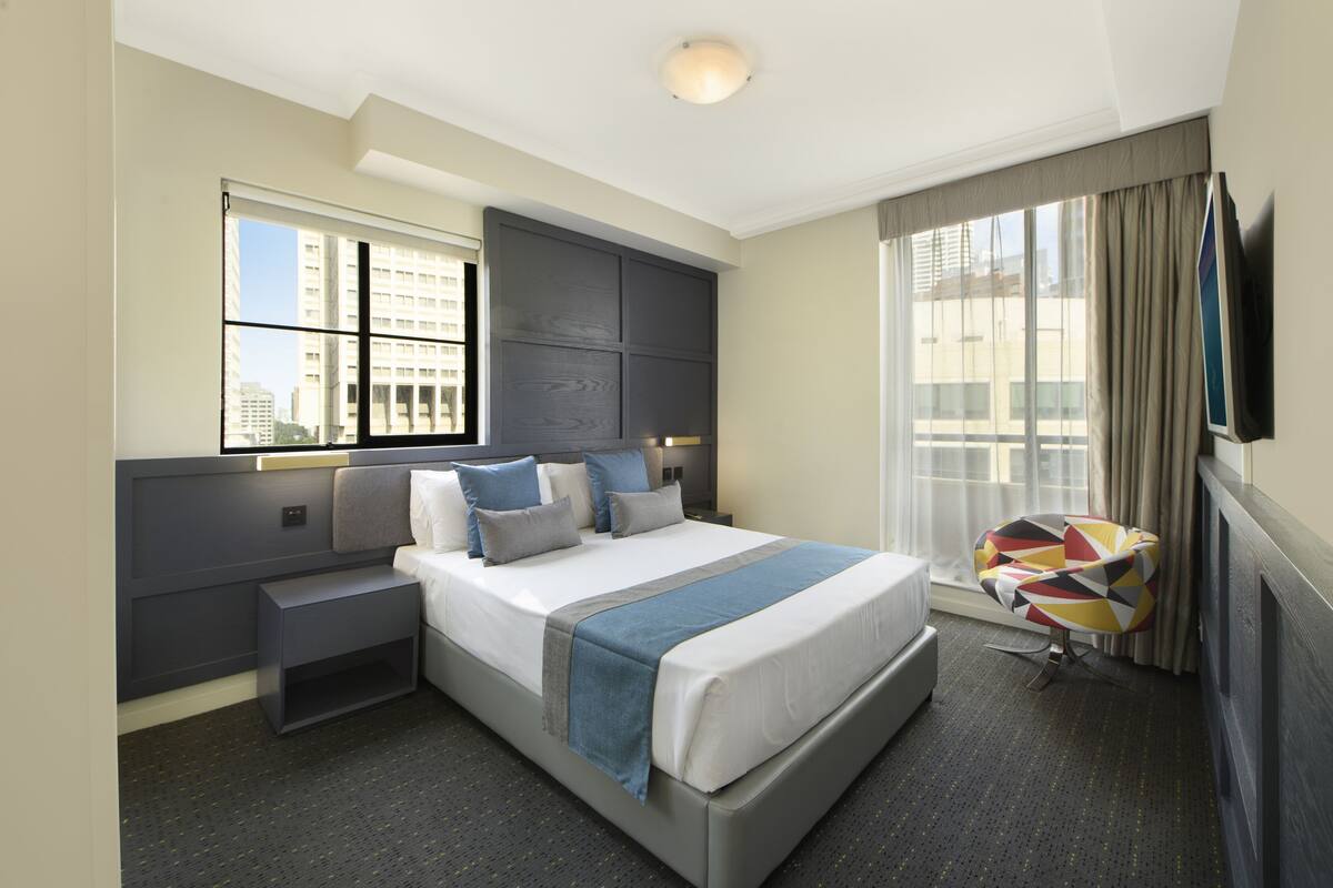 A hotel room with a large bed, a nightstand, a chair, a TV, and a window with curtains.