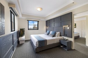 Una habitación de hotel moderna con una cama grande, un televisor de pantalla plana, un ventanal con vista a la ciudad y un cabecero oscuro.