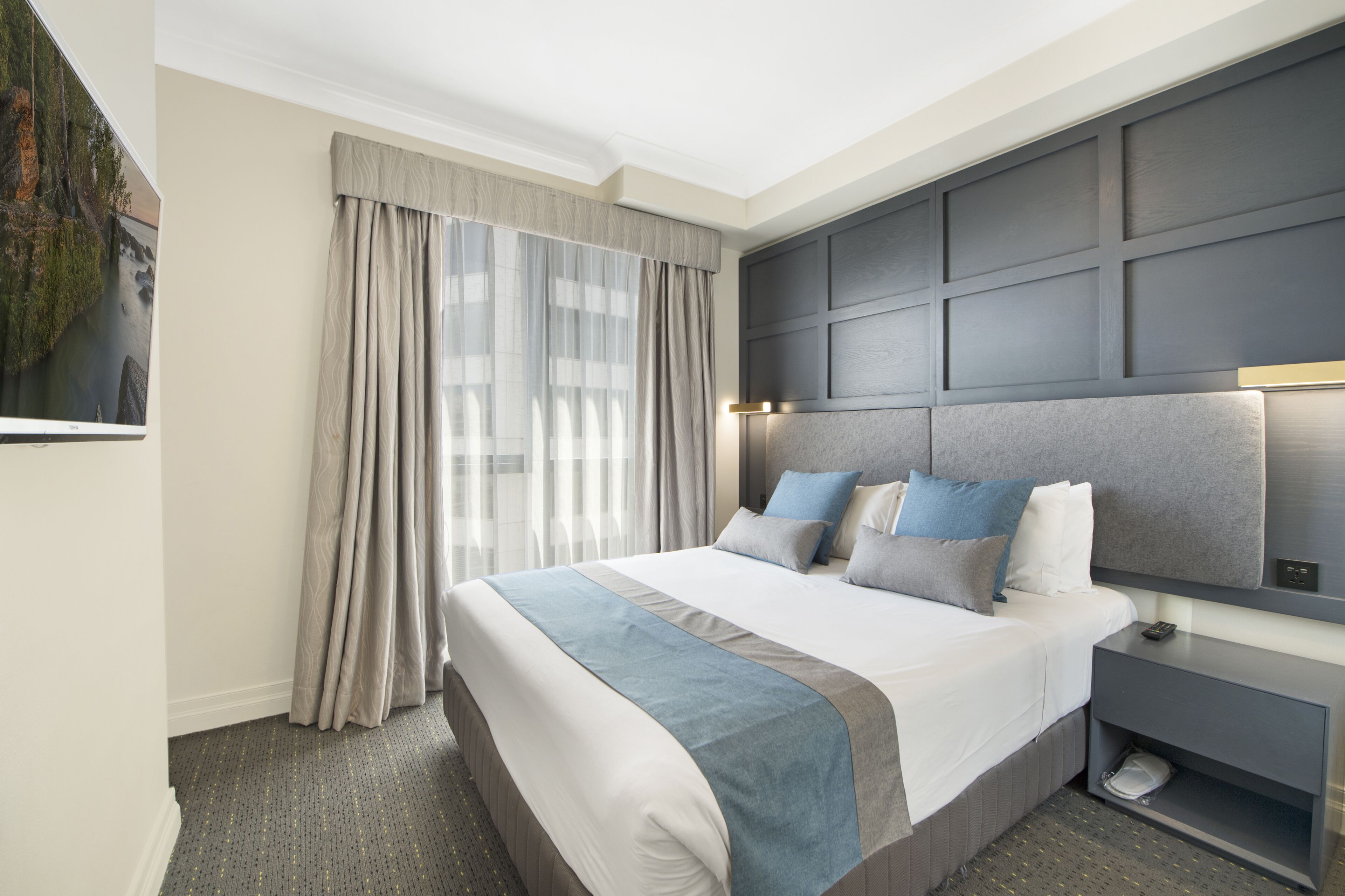 Hotelkamer met een groot bed, een hoofdeinde met ingebouwde verlichting, een nachtkastje met een lamp en uitzicht op een stadsbeeld.
