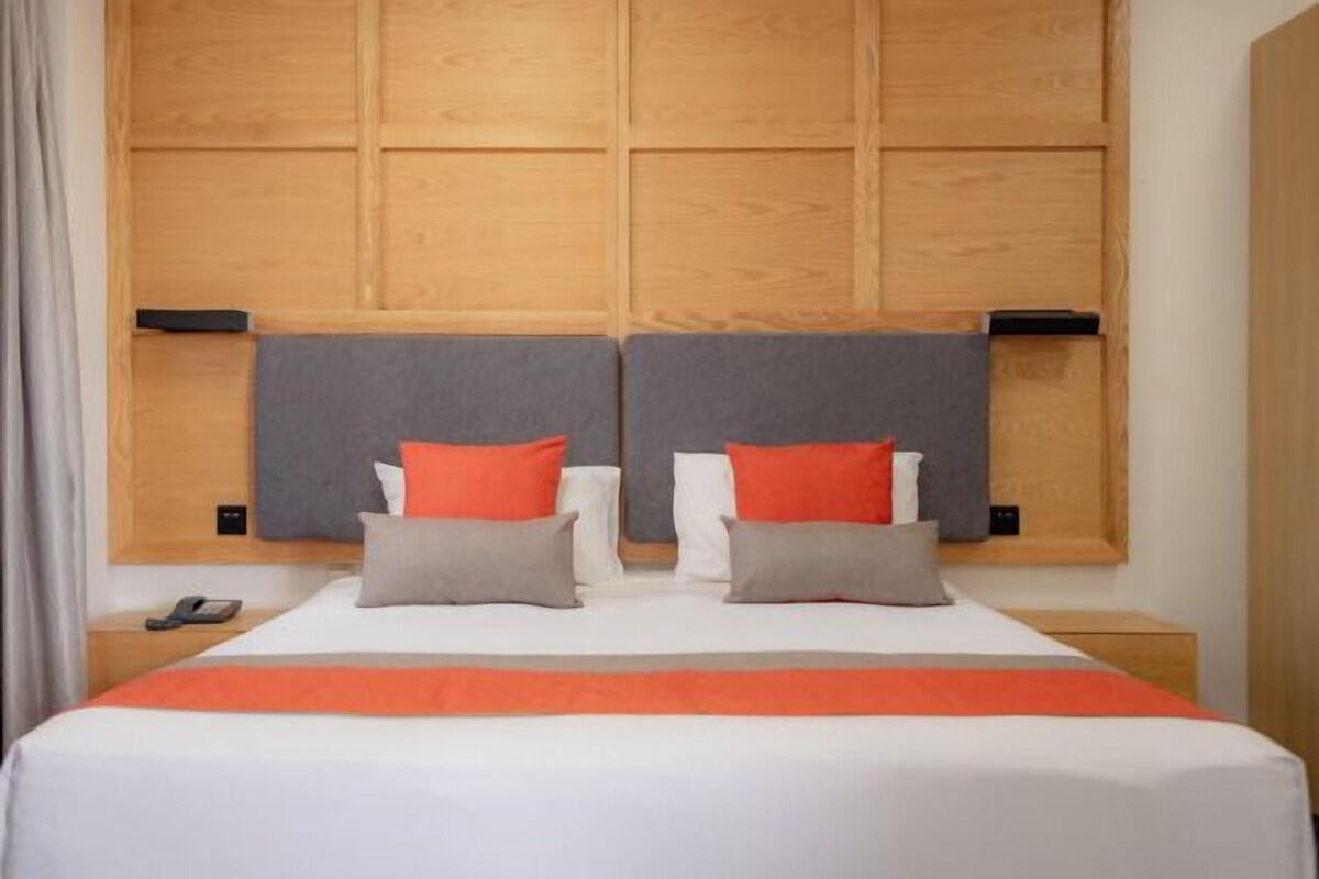 Una cama doble con cabecera de madera, dos almohadas, una manta y una mesita de noche con un teléfono.