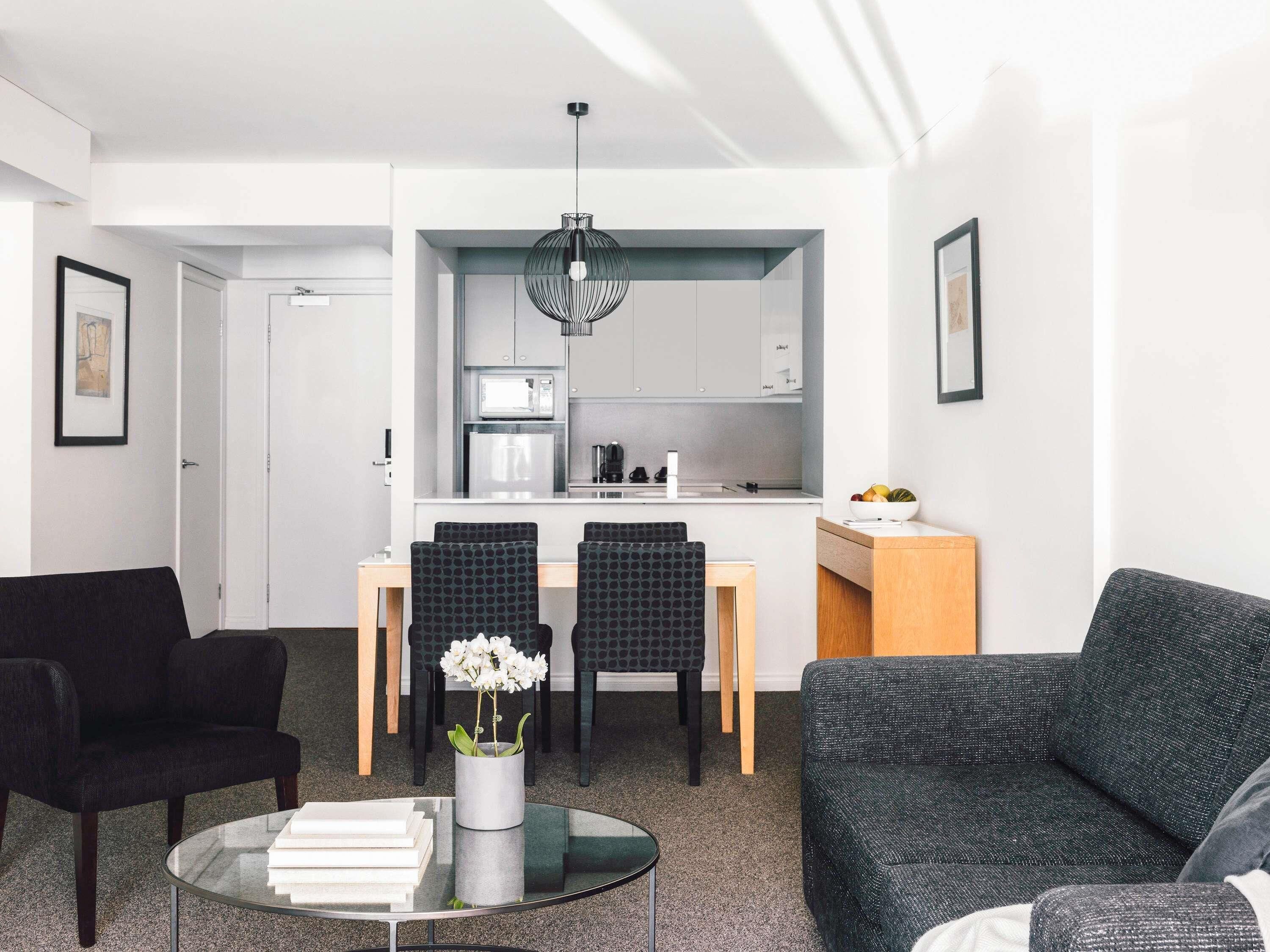 Ein modernes Wohnzimmer mit einem Glastisch, schwarzen und grauen Möbeln, einer Küchenzeile mit weißen Schränken und einer Pendelleuchte.