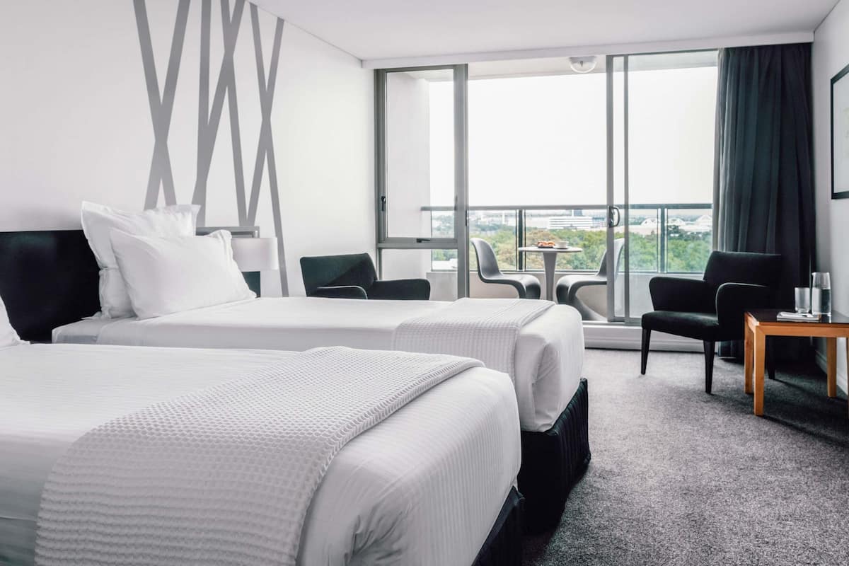 Une chambre d’hôtel avec deux lits, une petite table, deux chaises, une grande fenêtre donnant sur un paysage agréable et un mur orné d’un motif géométrique.