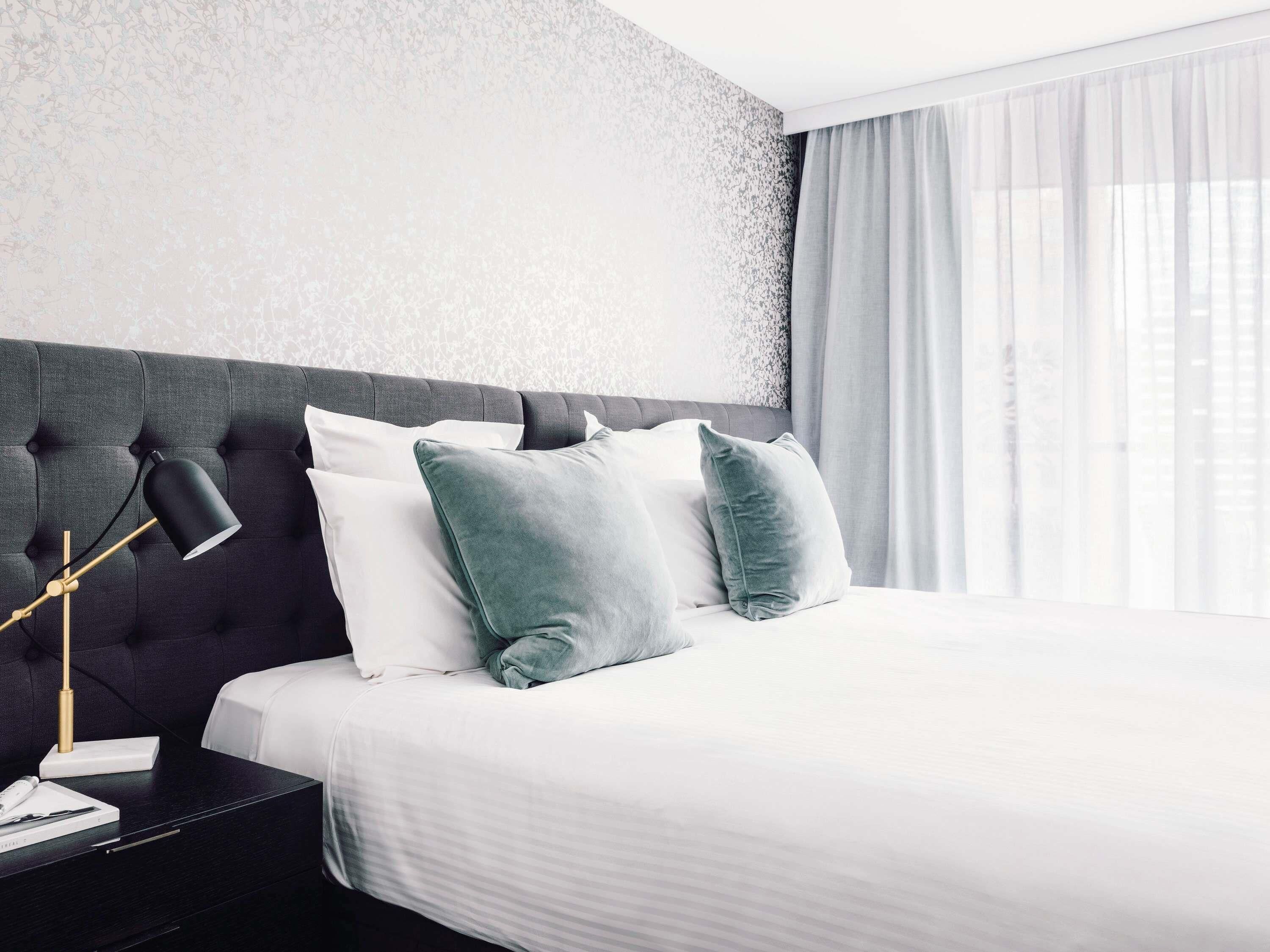 Ett modernt hotellrum med en stoppad sänggavel, en sänglampa och ett stort fönster med tunna, genomskinliga gardiner.