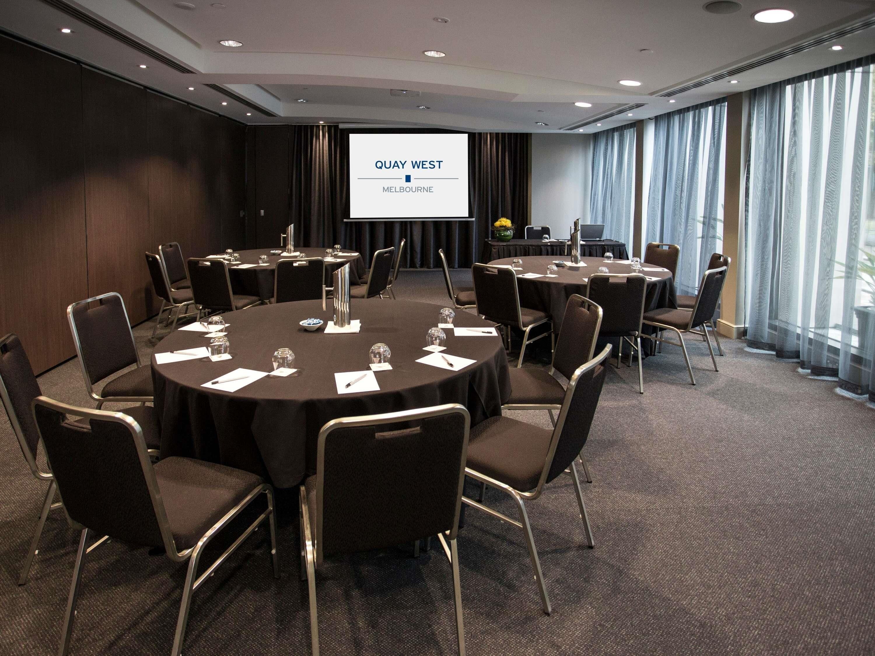 Un salón de conferencias con mesas redondas, sillas, una pantalla de proyector mostrando 'QUAY WEST MELBOURNE' y amplias ventanas con cortinas.