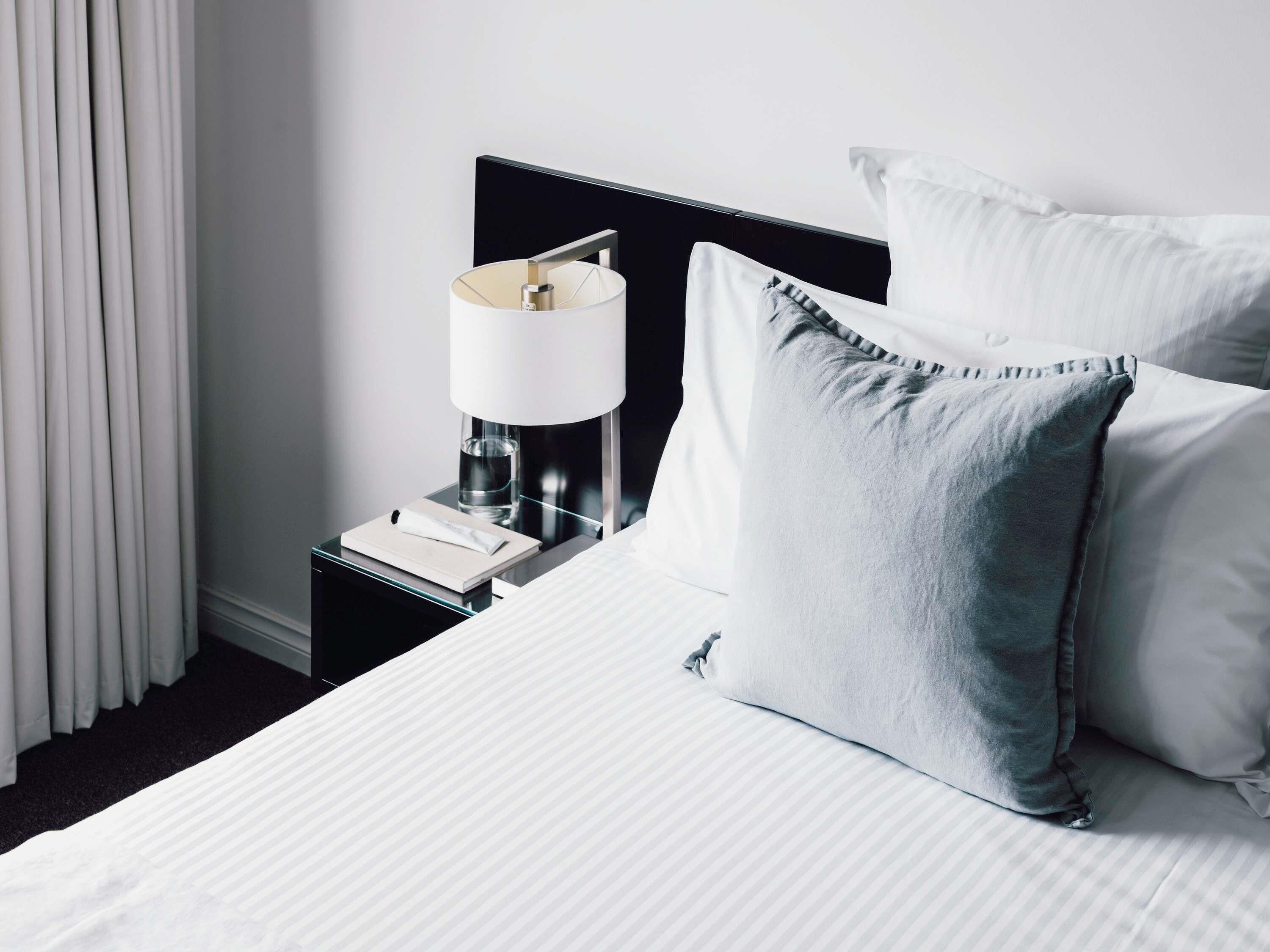 A neatly made bed with white bedding and a grey pillow, a bedside table with a lamp and a book, and a window with white curtains.