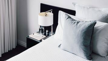 A neatly made bed with white bedding and a grey pillow, a bedside table with a lamp and a book, and a window with white curtains.