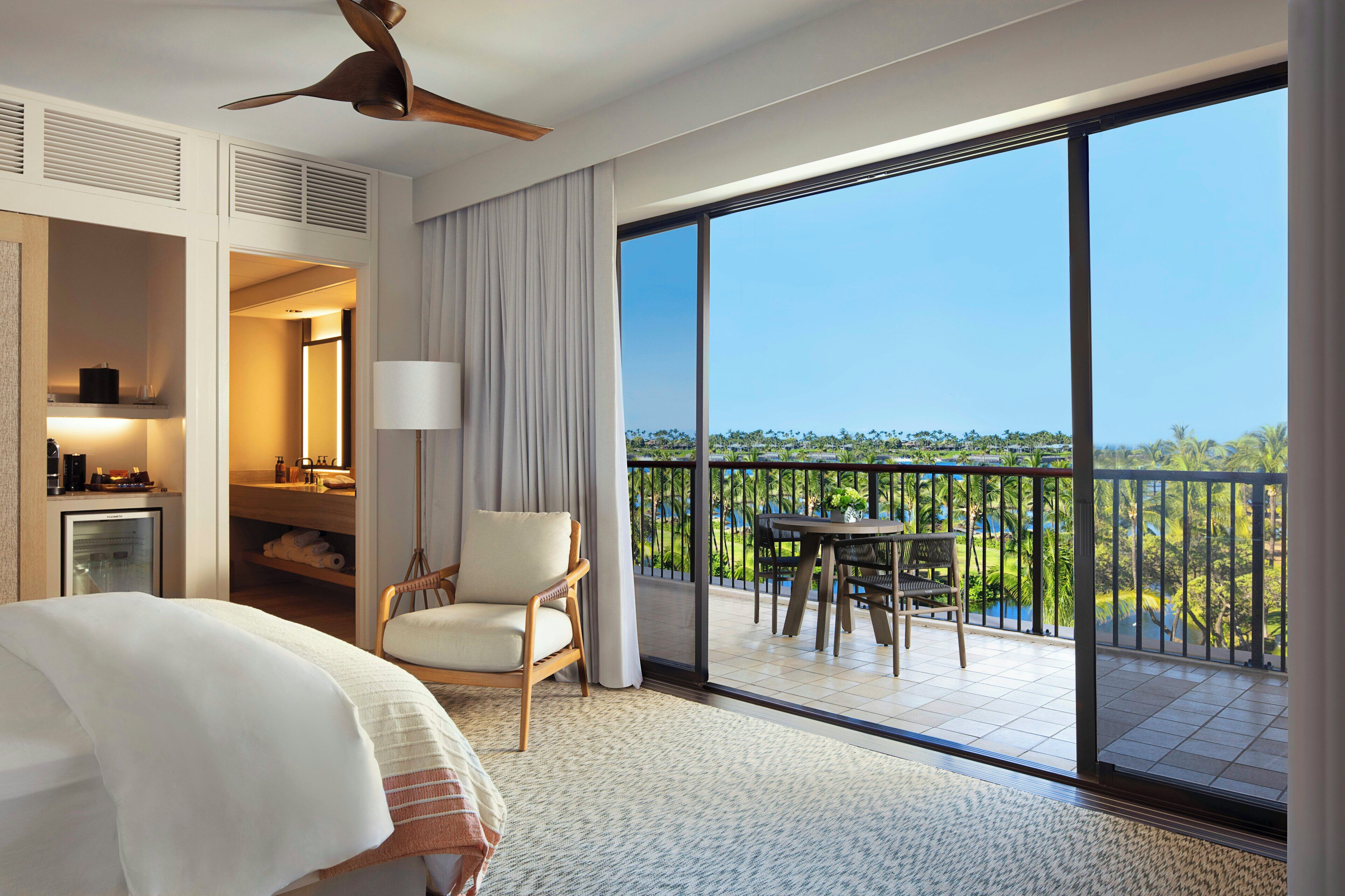 A hotel room with a bed, a chair, a table, a balcony with a view, and a ceiling fan.