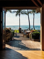 An outdoor patio with wooden flooring, a table with chairs, and a view of the ocean with palm trees.