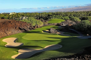 A golf course with bunkers, a flag, and a view of a coastal area with buildings in the background.