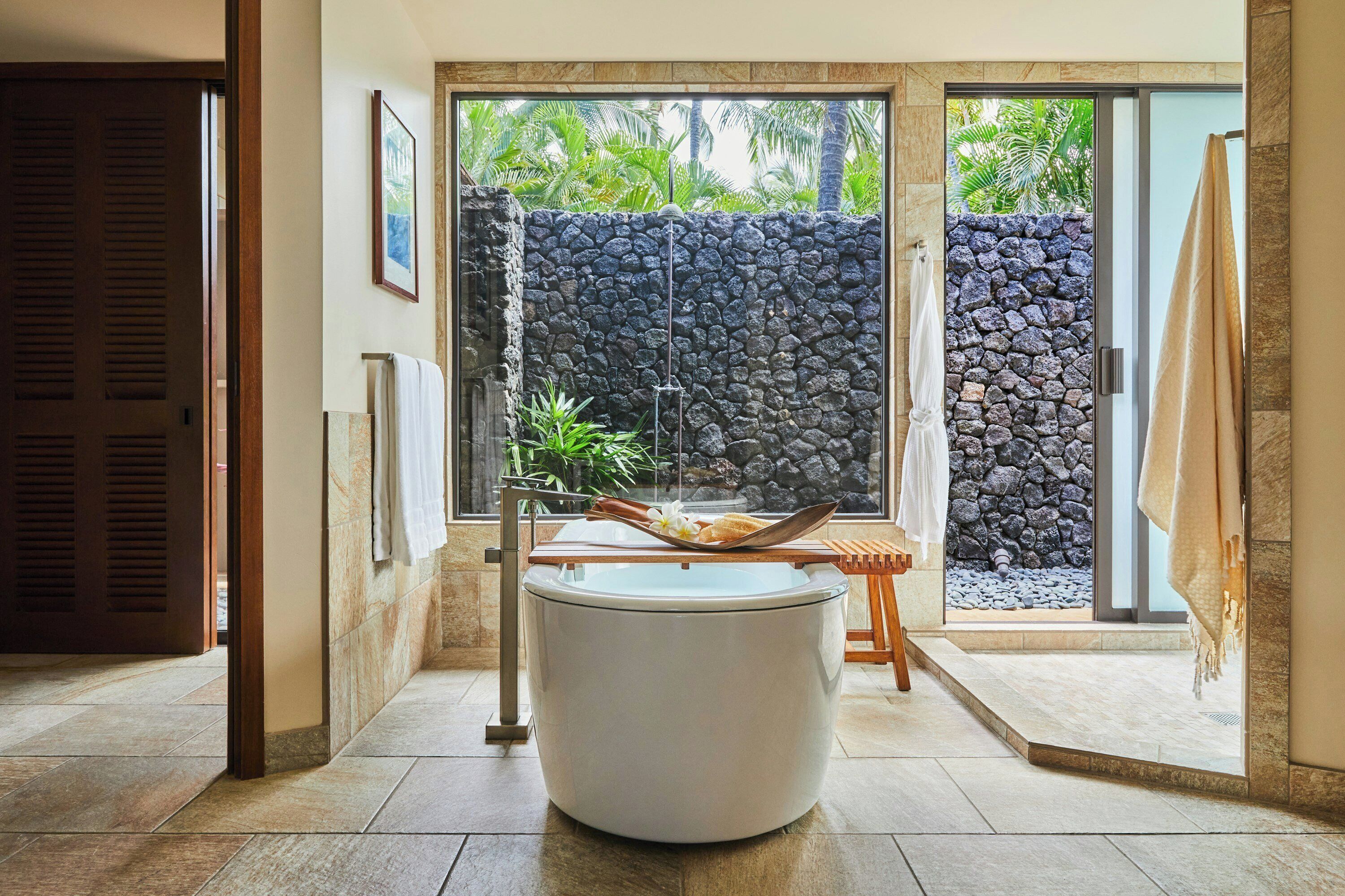 A bathroom with a large bathtub, a stone wall, and a view of greenery.