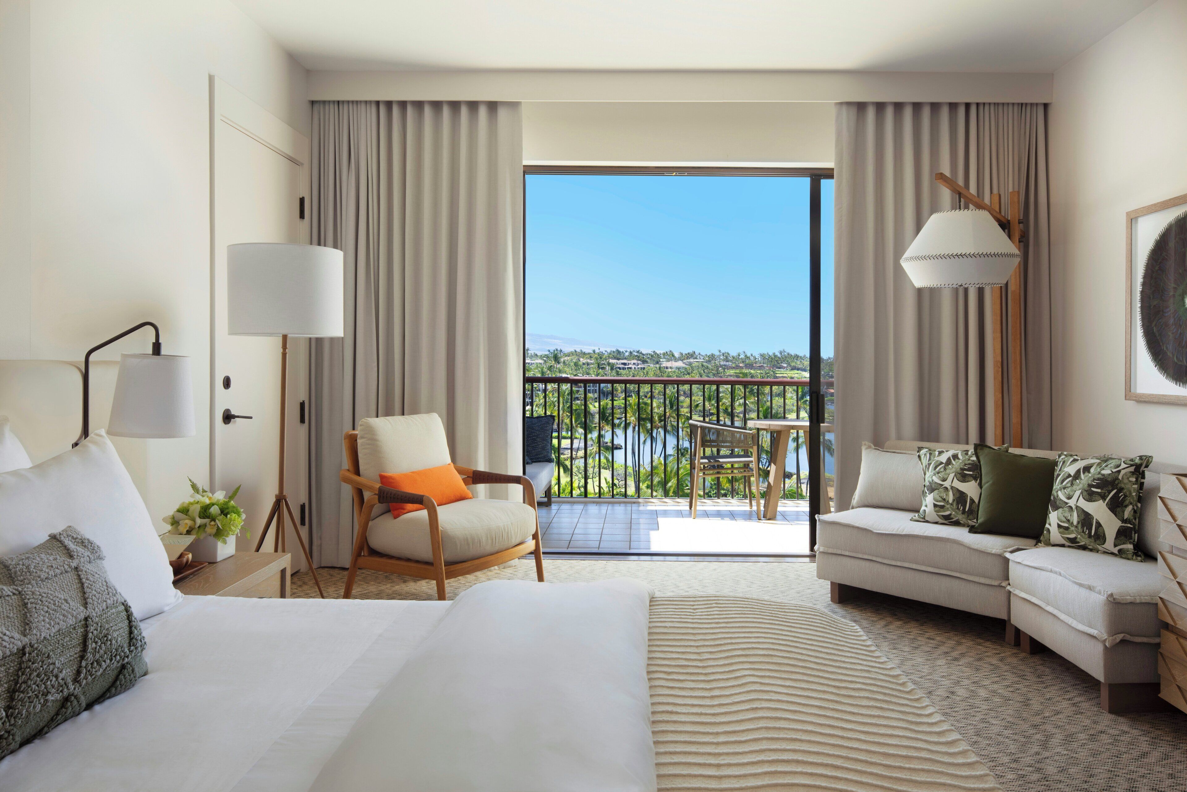 Una habitación de hotel con cama, sofá, silla, escritorio, lámpara, ventana con cortinas y balcón con vistas.