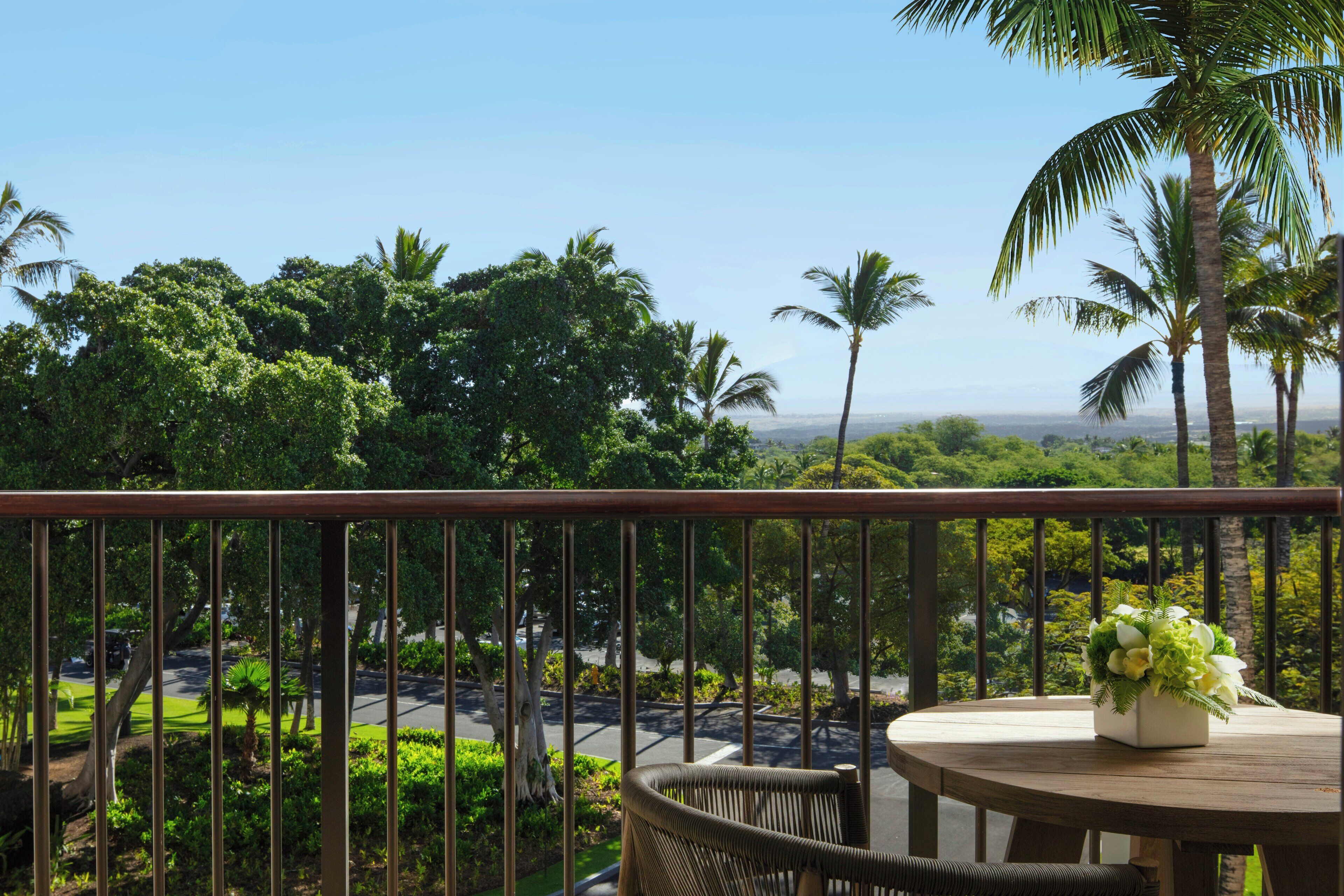 A balcony with a table and chairs, overlooking a lush green landscape with palm trees and a view of the ocean.