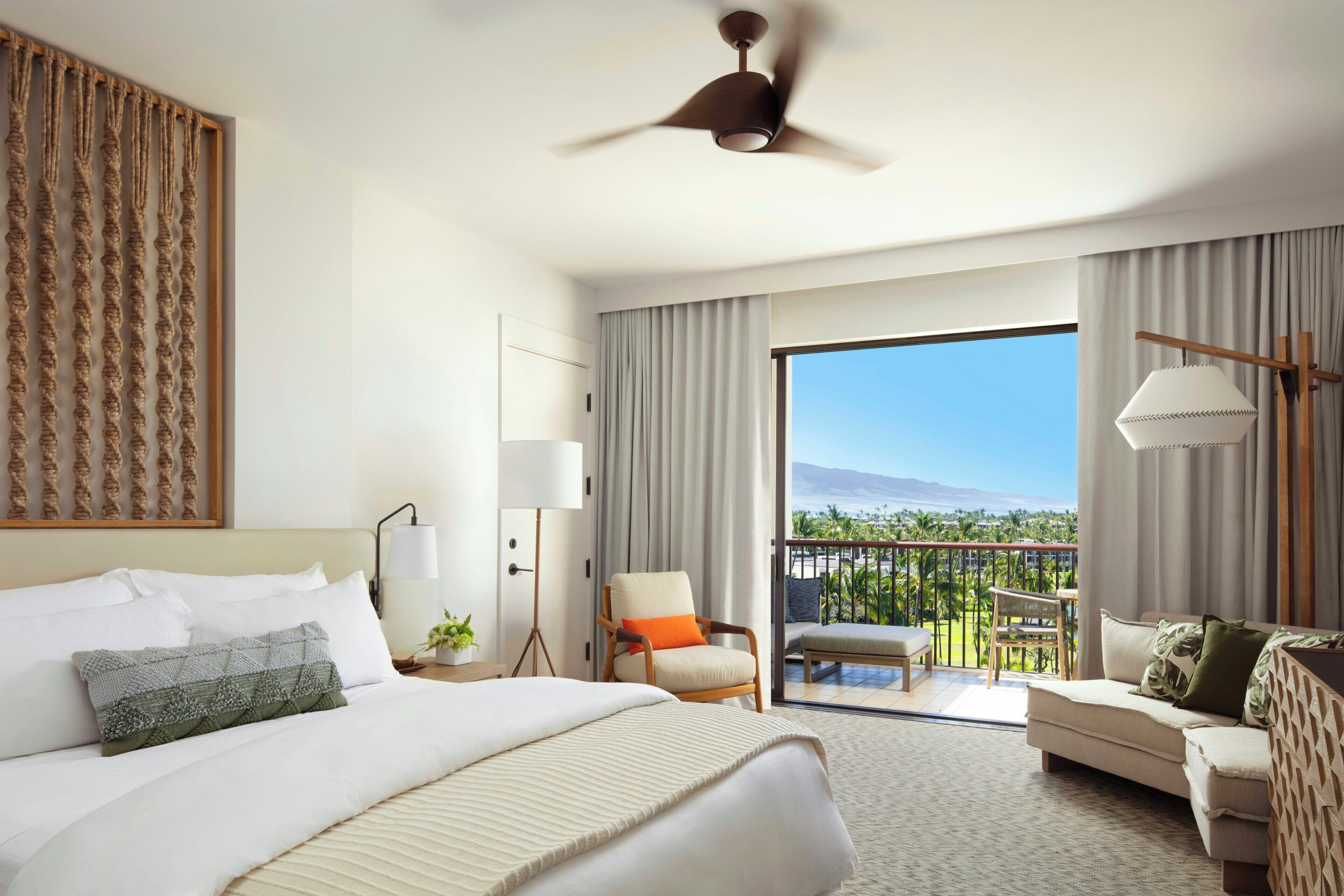 A hotel room with a bed, a chair, a sofa, a balcony with a view, and a ceiling fan.