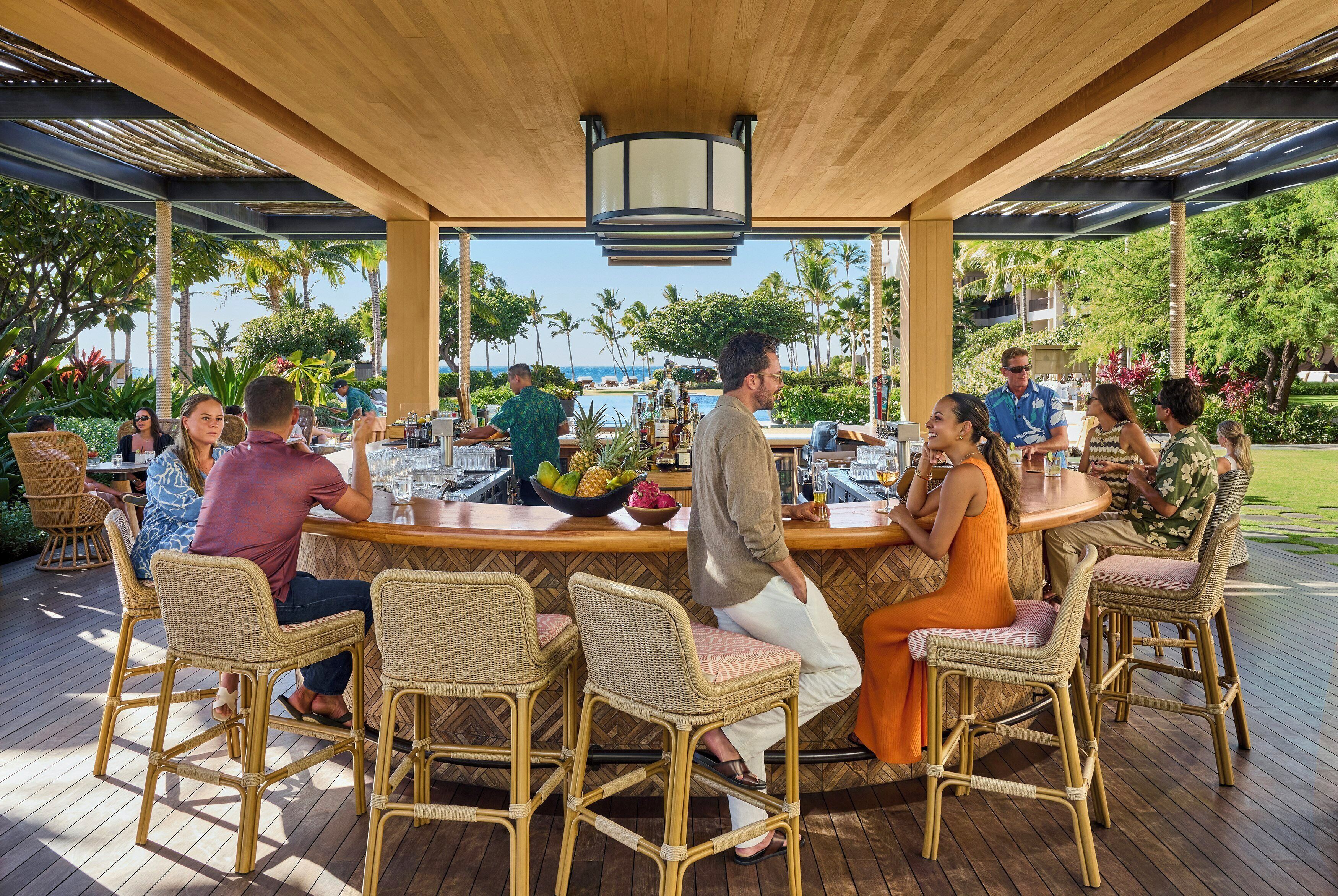 An outdoor bar area with wicker chairs and a wooden ceiling, surrounded by lush greenery and a view of the ocean.