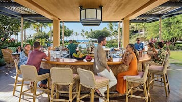 An outdoor bar area with wicker chairs and a wooden ceiling, surrounded by lush greenery and a view of the ocean.