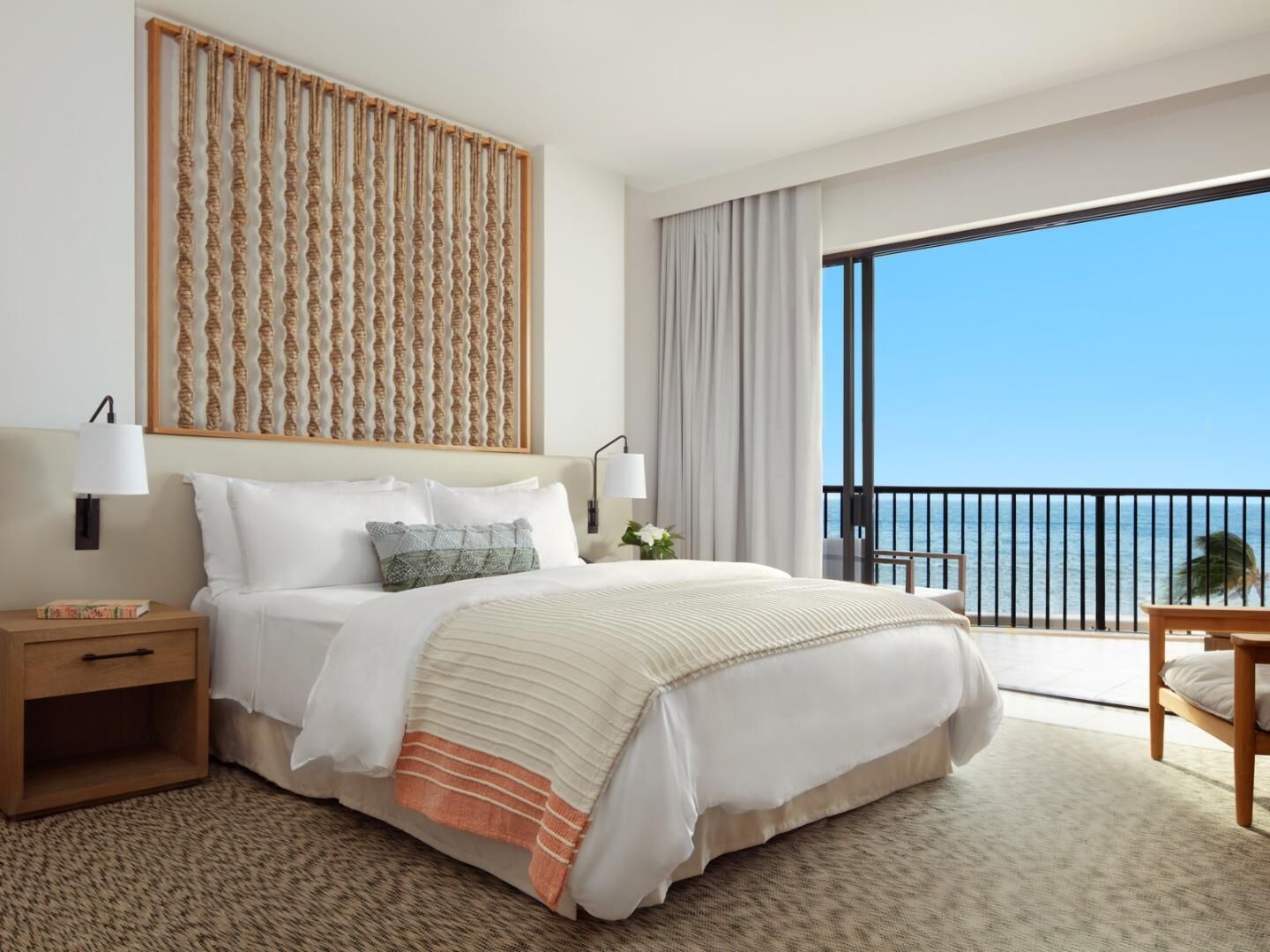 A hotel room with a bed, bedside tables, a view of the ocean, and a balcony.