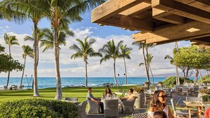 A tropical outdoor dining area with wicker furniture, palm trees, and a view of the ocean. - Mauna Lani, Auberge Collection (Kamuela)